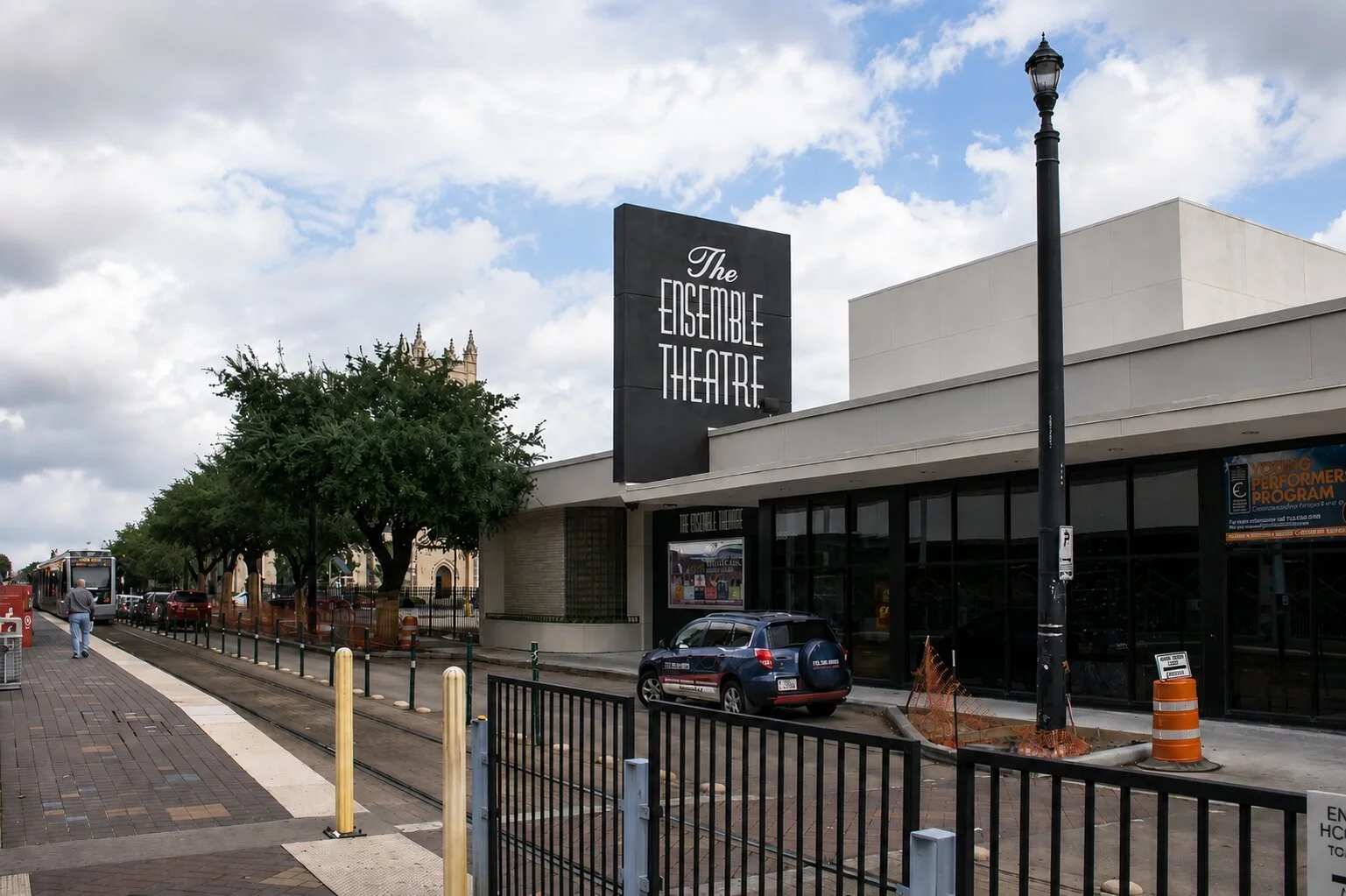 Street view of The Ensemble Theatre in Houston with a tall exterior sign, nearby light rail tracks, glass facade, and theater entrance.