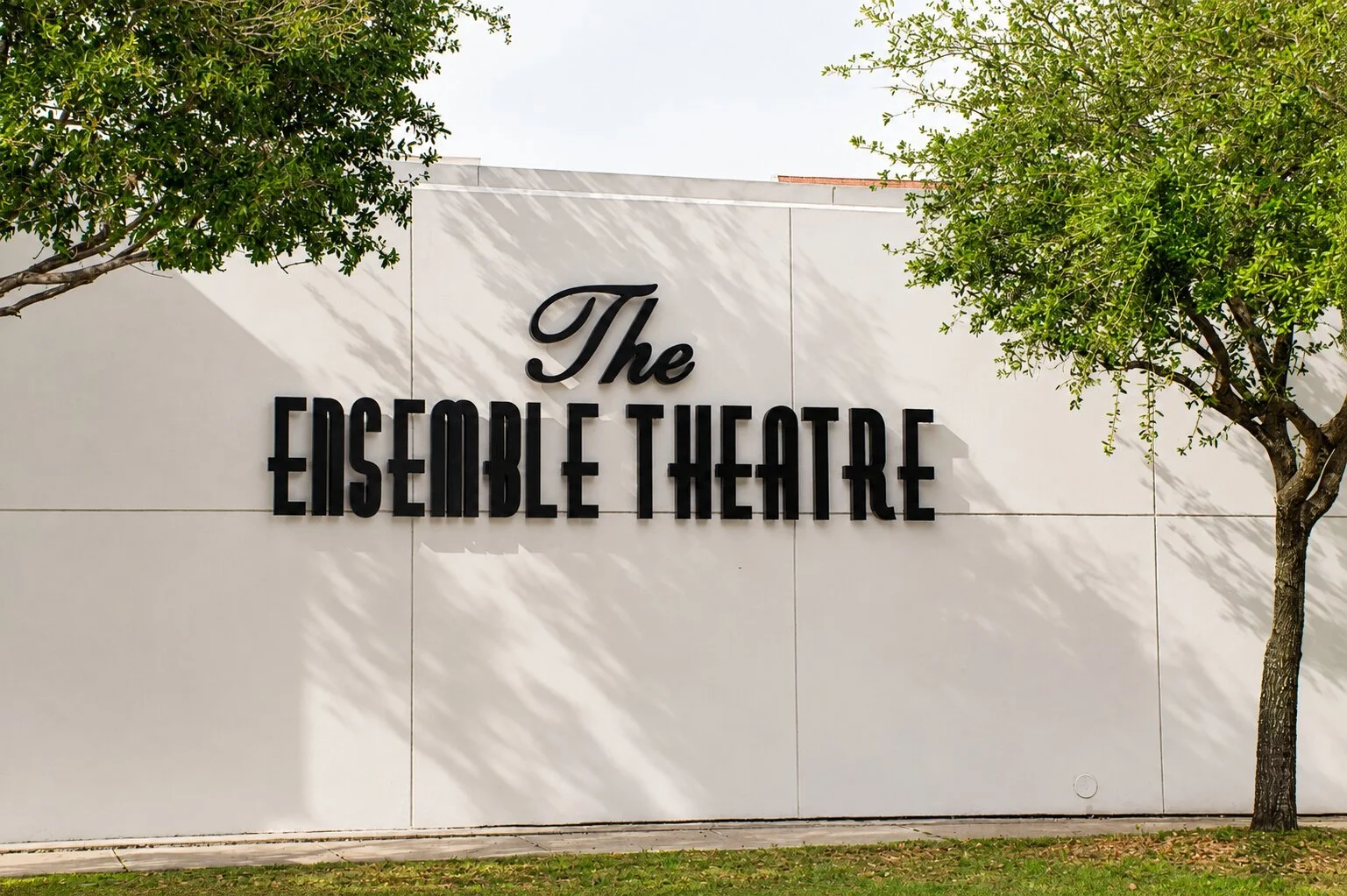The Ensemble Theatre in Houston with large black dimensional letters mounted on a white exterior wall between mature trees.