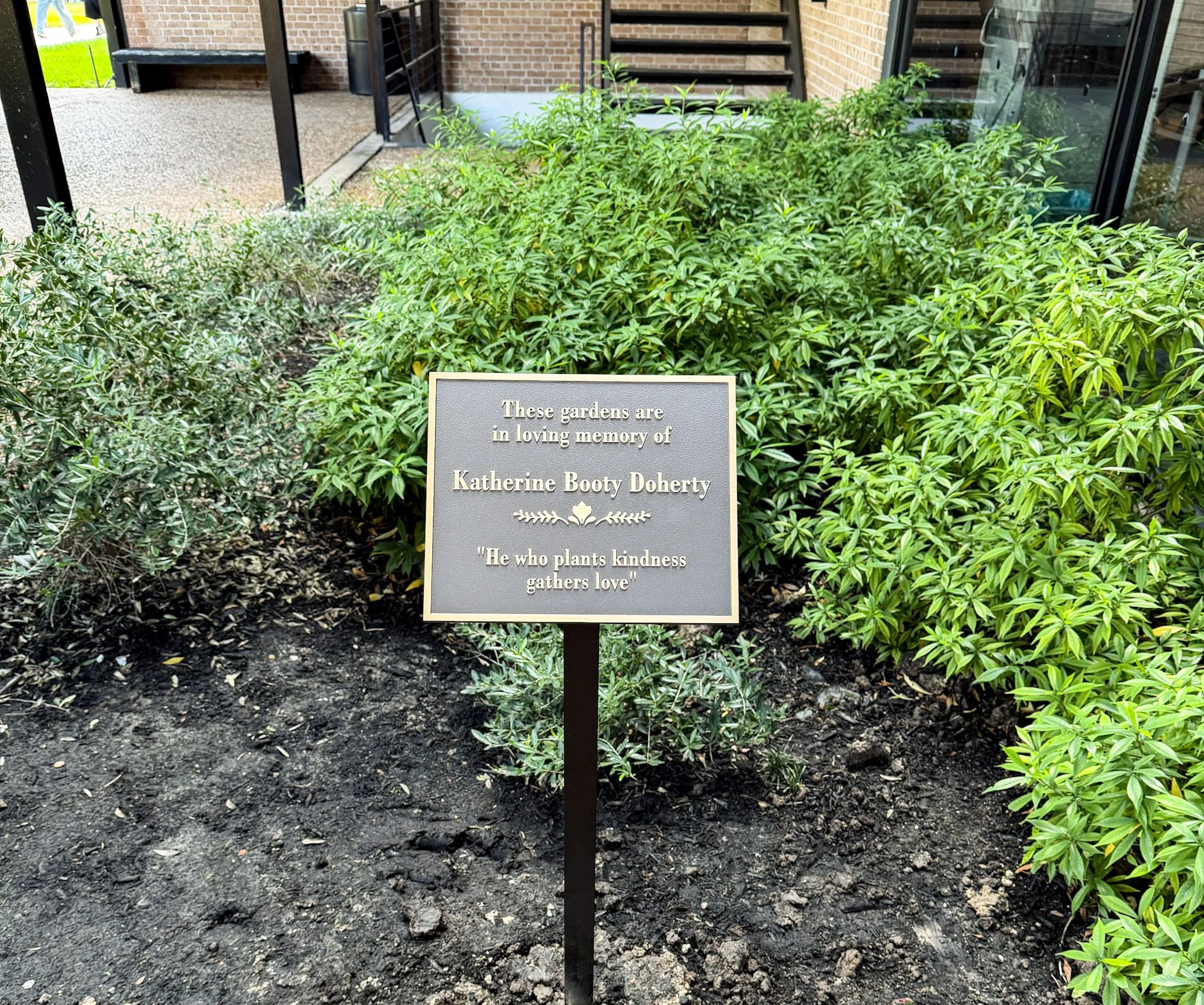 Bronze garden memorial plaque honoring Katherine Booty Doherty, installed on a post among landscaped plants and shrubs.