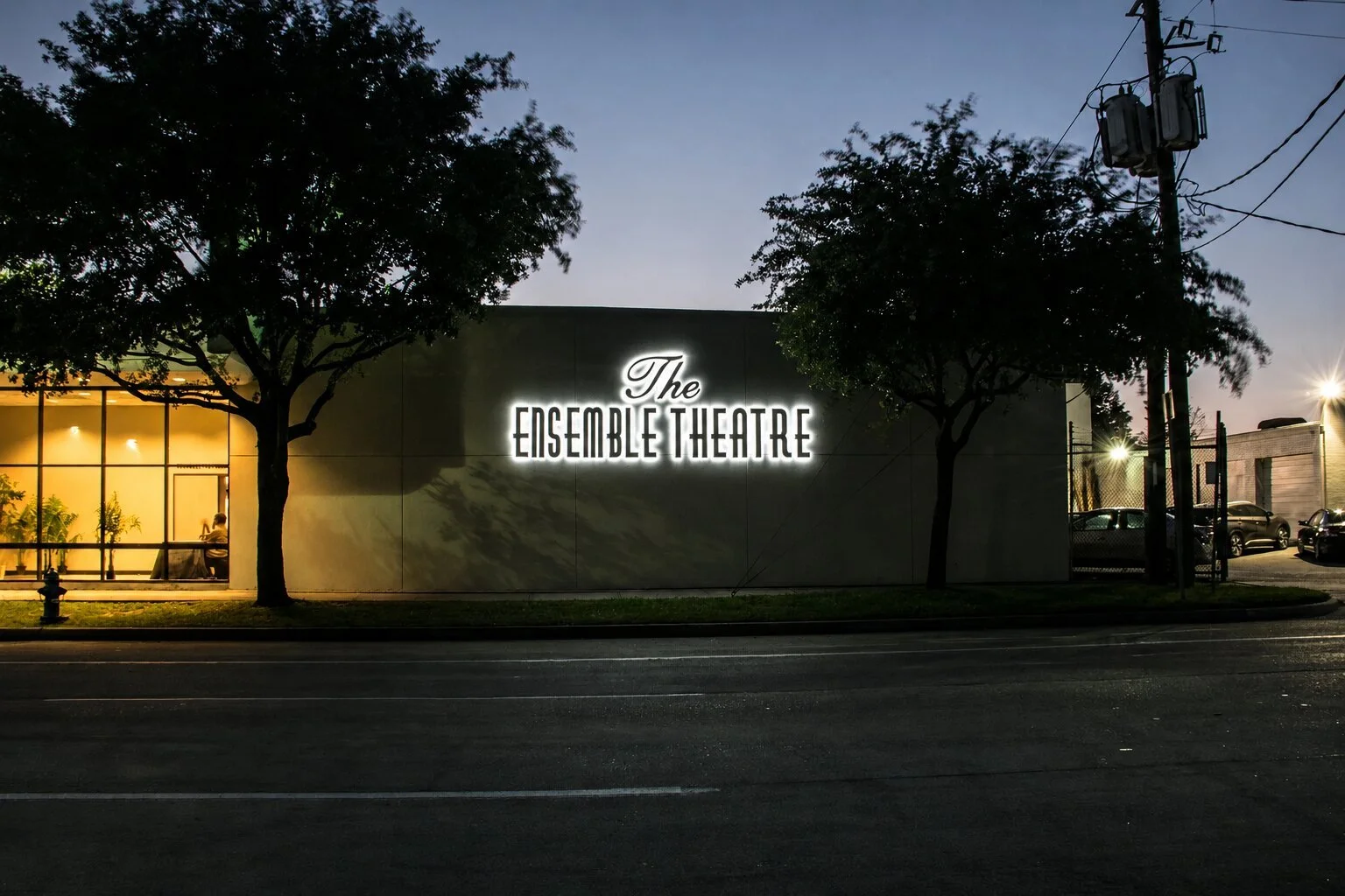 The Ensemble Theatre in Houston at dusk with an illuminated exterior wall sign visible between trees across the street.