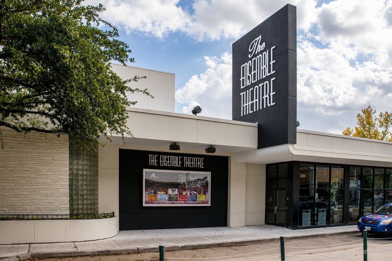 The Ensemble Theatre in Houston showing the main entrance, tall exterior building sign, framed display case, glass doors, and surrounding facade.