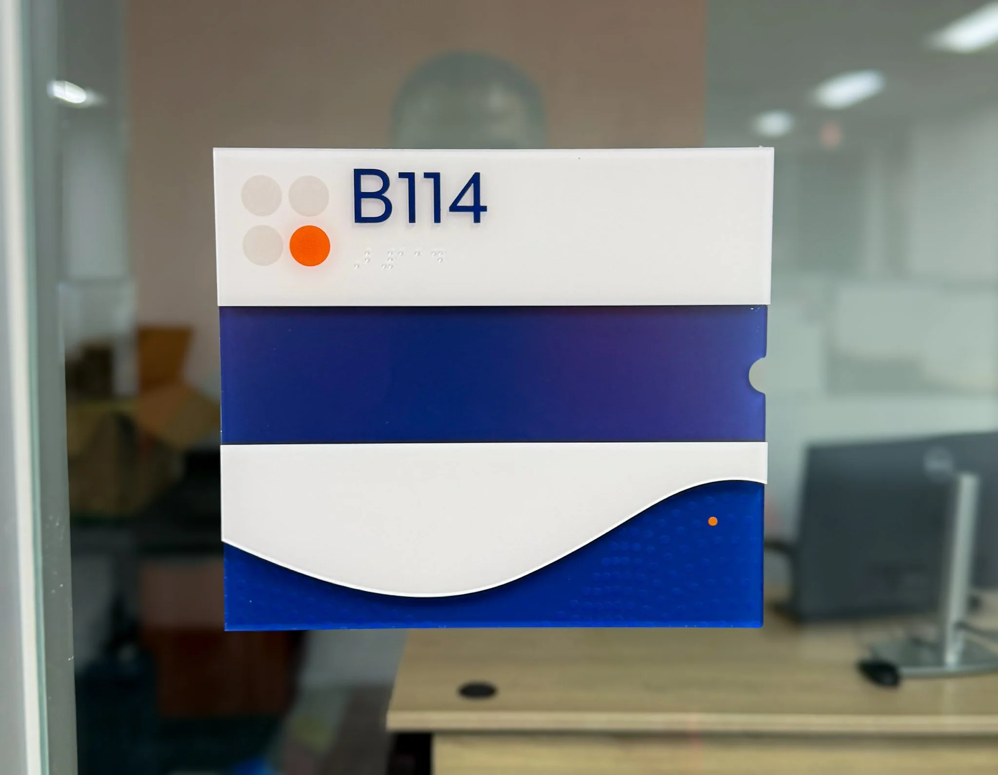 ADA room sign for B114 with raised lettering, Braille, and blue and orange graphics.