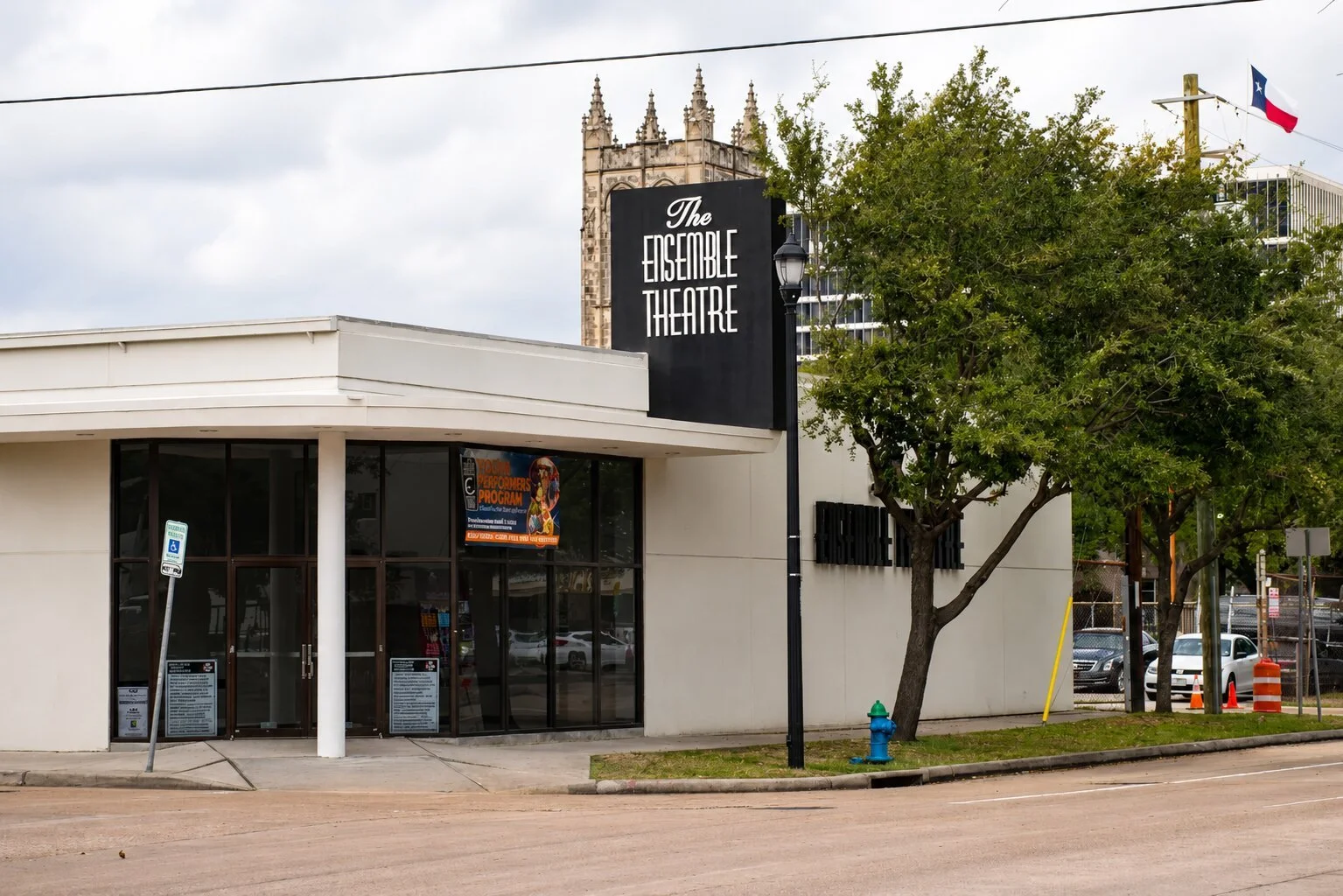 Street view of The Ensemble Theatre in Houston with a tall exterior sign, glass entrance, mature trees, and surrounding urban buildings.
