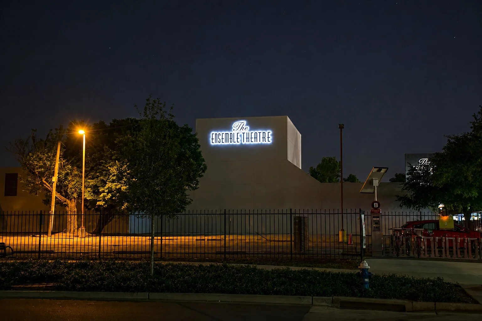 Nighttime view of The Ensemble Theatre in Houston with an illuminated exterior building sign visible above the surrounding trees and fence.