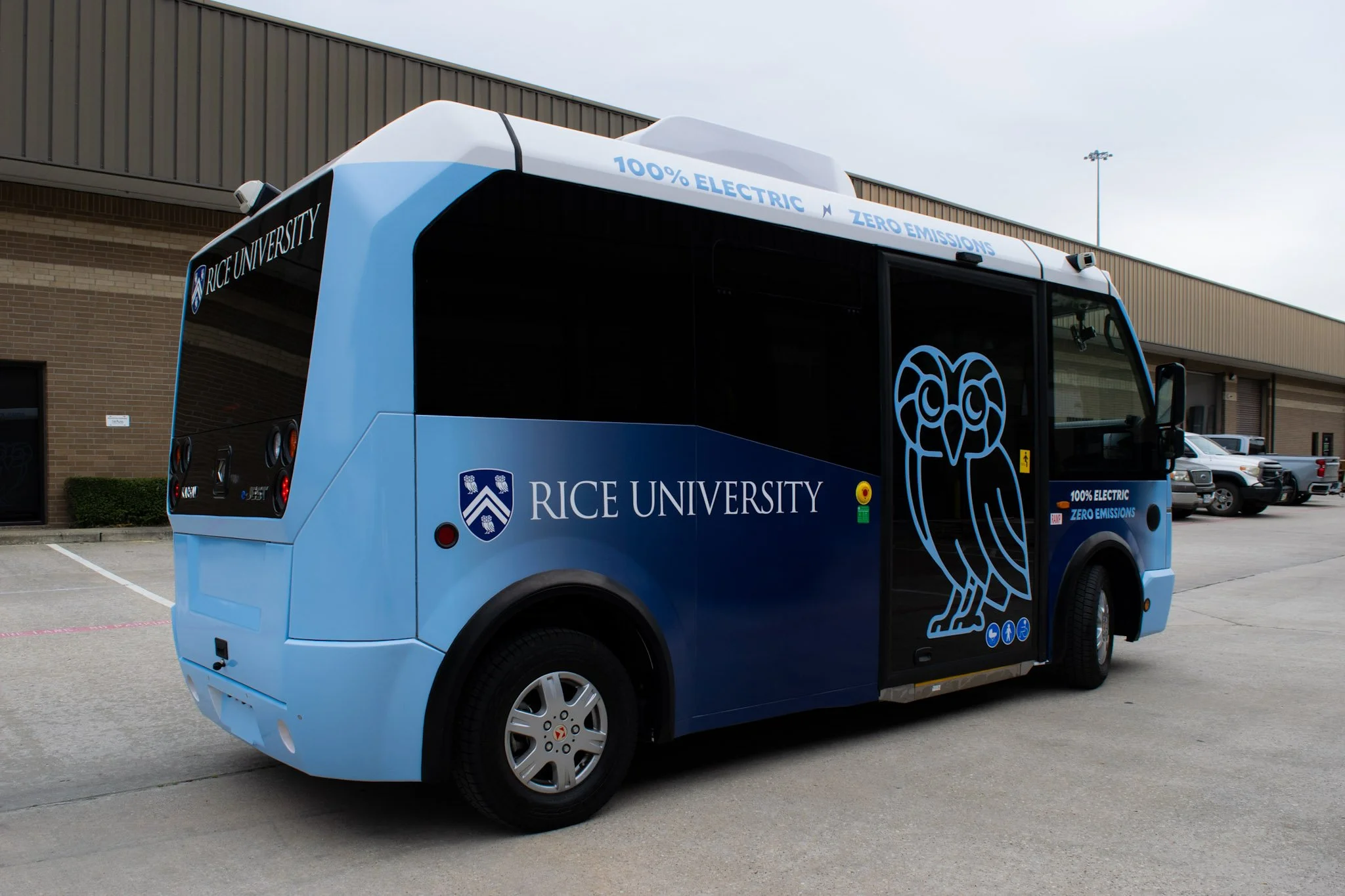 Houston electric shuttle bus wrap for Rice University featuring blue gradient branding, owl graphic, and “100% Electric Zero Emissions” messaging parked outside facility.