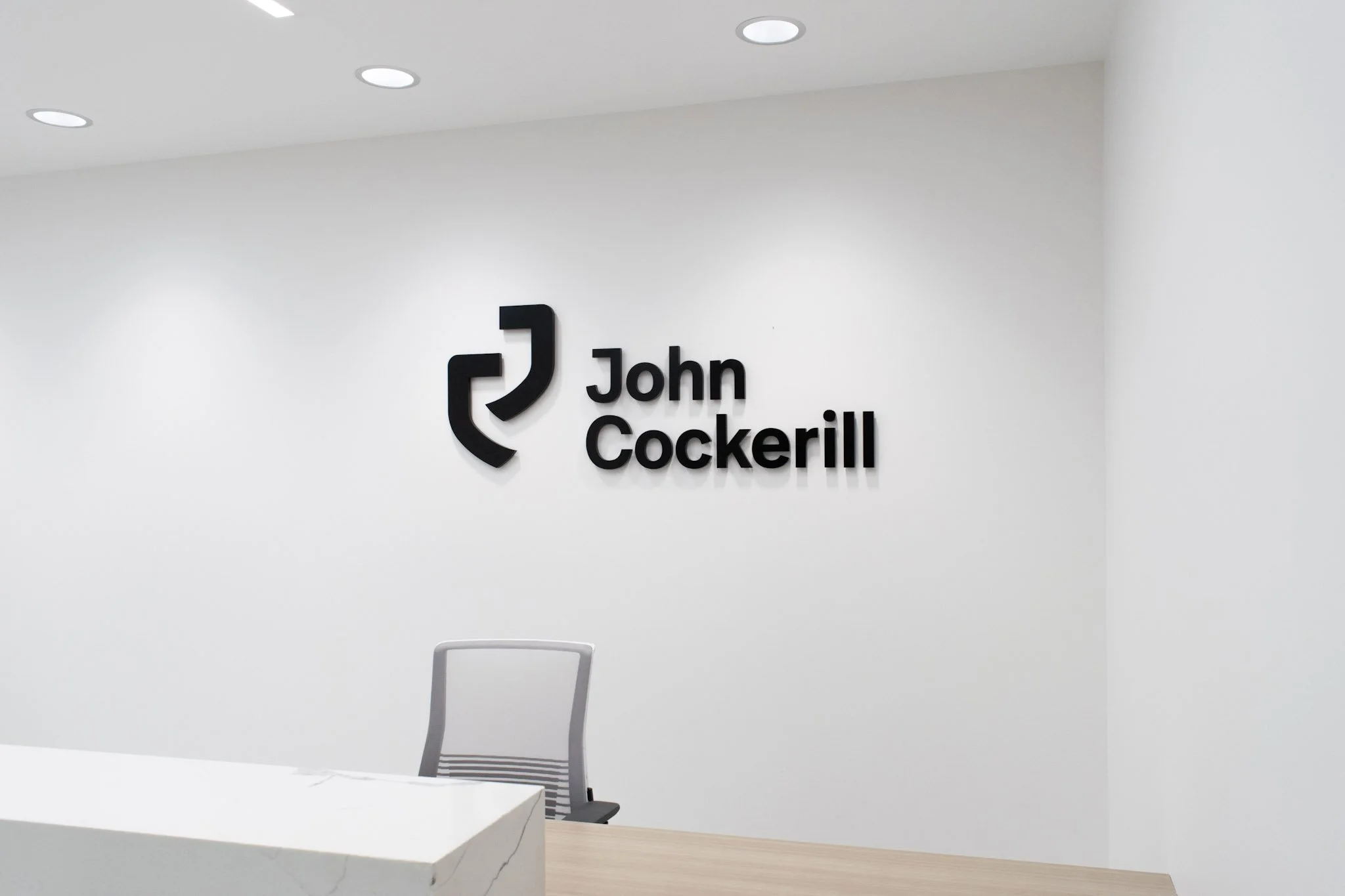 Black dimensional logo and lettering for John Cockerill installed on a white reception wall.