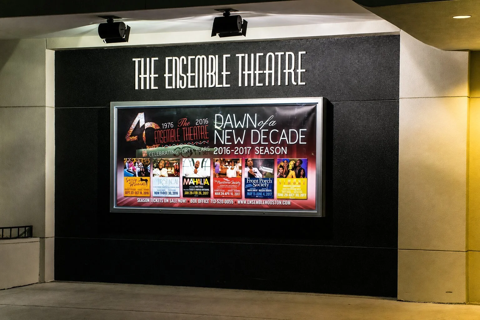 The Ensemble Theatre in Houston with exterior lettering above a framed season poster display on a black wall illuminated by overhead lights.