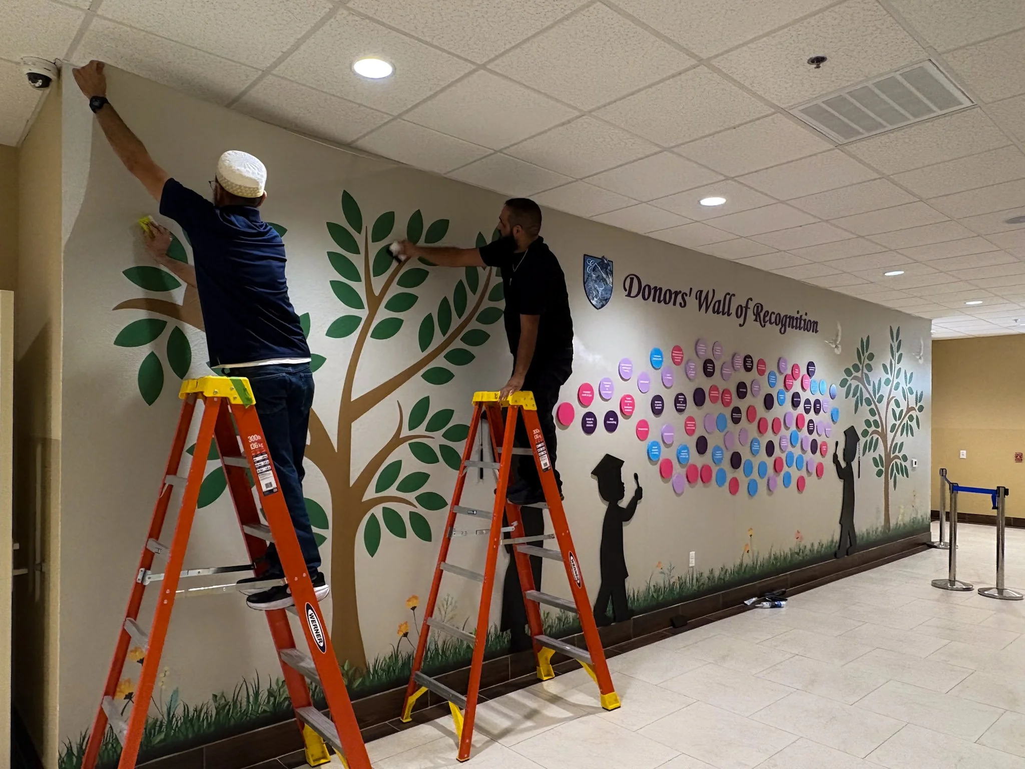 Installers applying donor recognition wall graphics and tree designs along a commercial interior hallway.