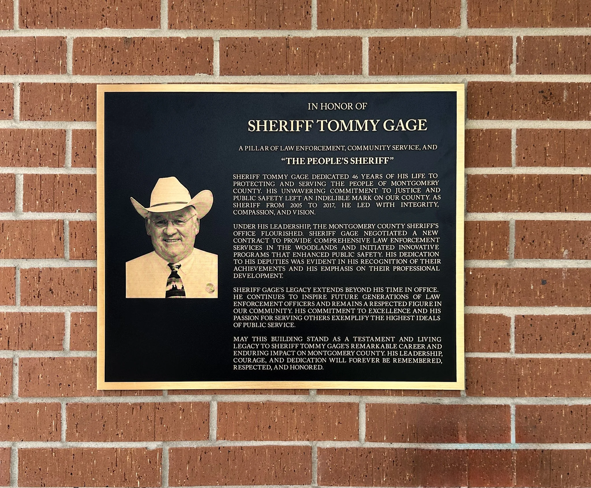 Custom commemorative plaque honoring Sheriff Tommy Gage, featuring a portrait and raised lettering mounted on an exterior brick wall.