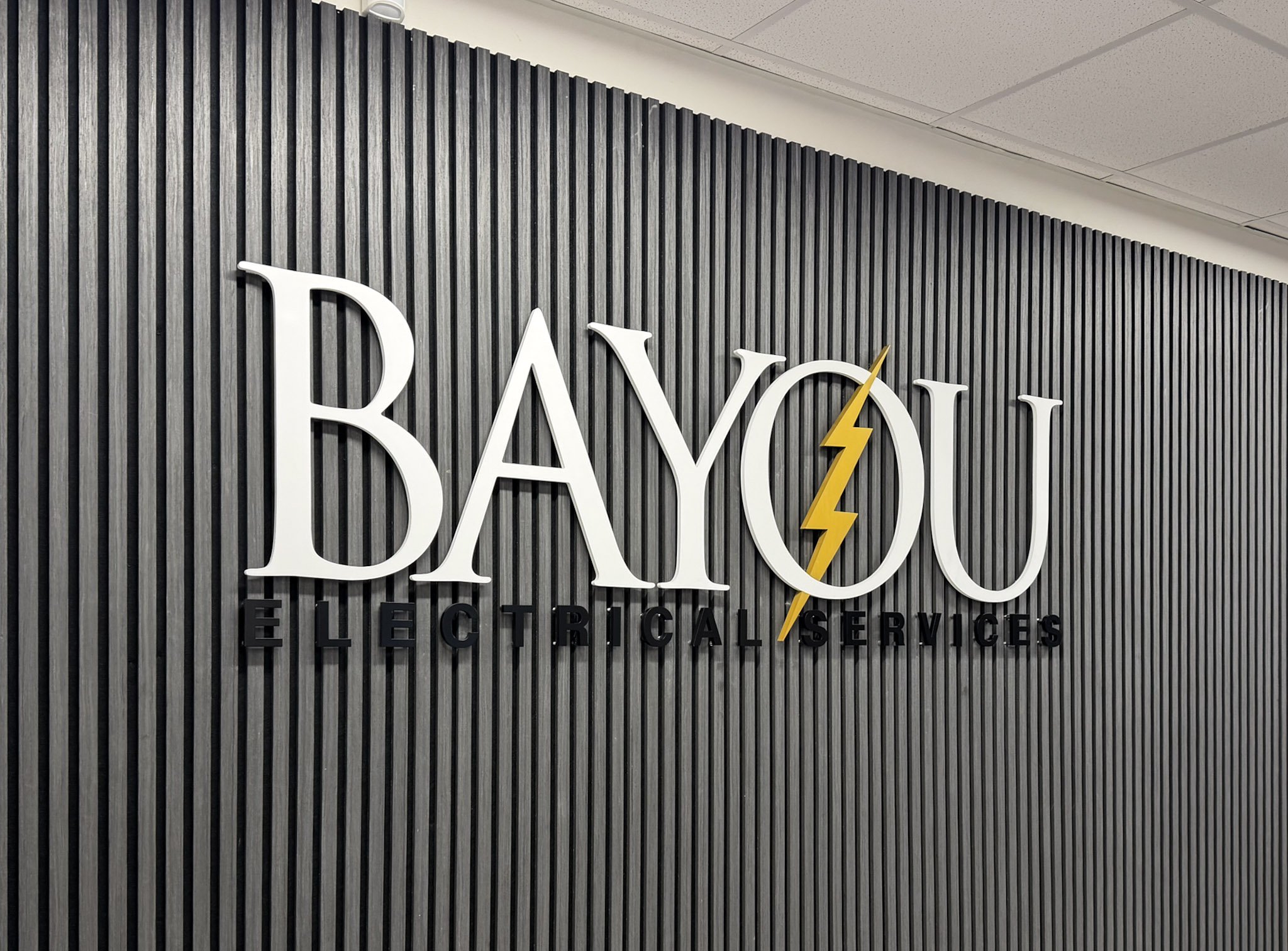 Interior dimensional logo sign for Bayou Electrical Services in Houston with white lettering and yellow lightning bolt on textured wall.