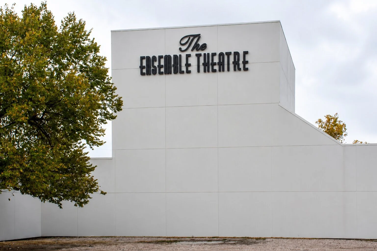 The Ensemble Theatre in Houston with black dimensional exterior letters mounted high on a white building facade beside a large tree.