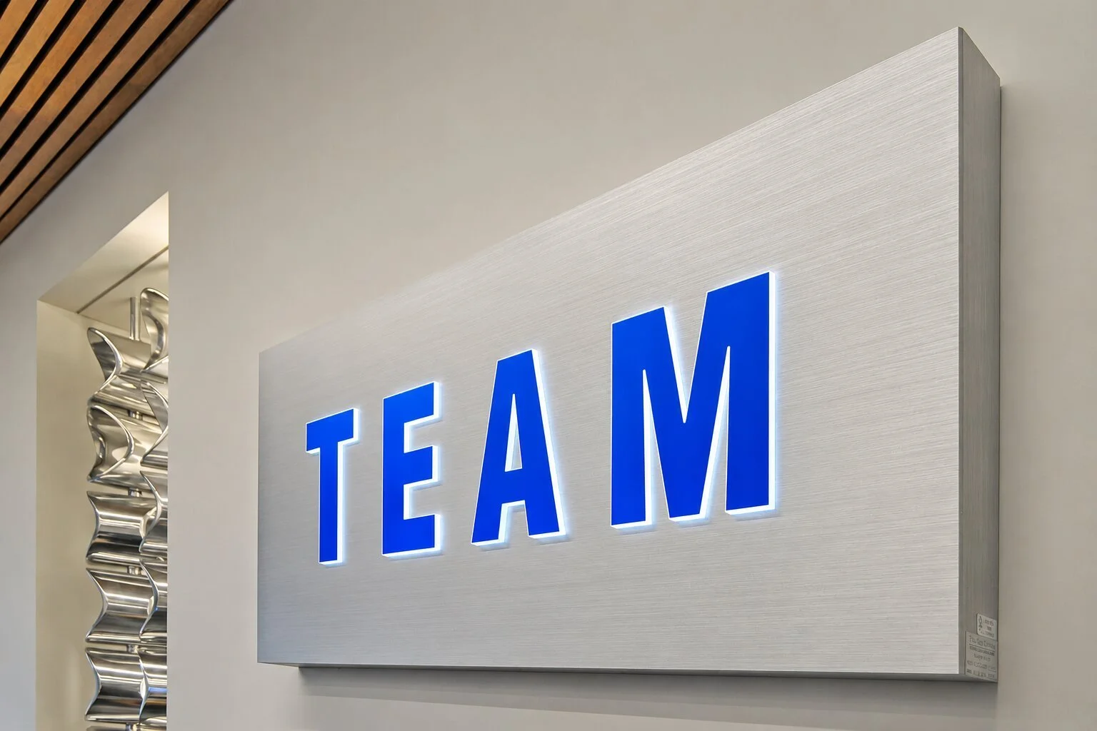 Backlit blue acrylic lobby logo sign with halo illumination mounted on a brushed metal backer panel in a modern corporate office reception area.