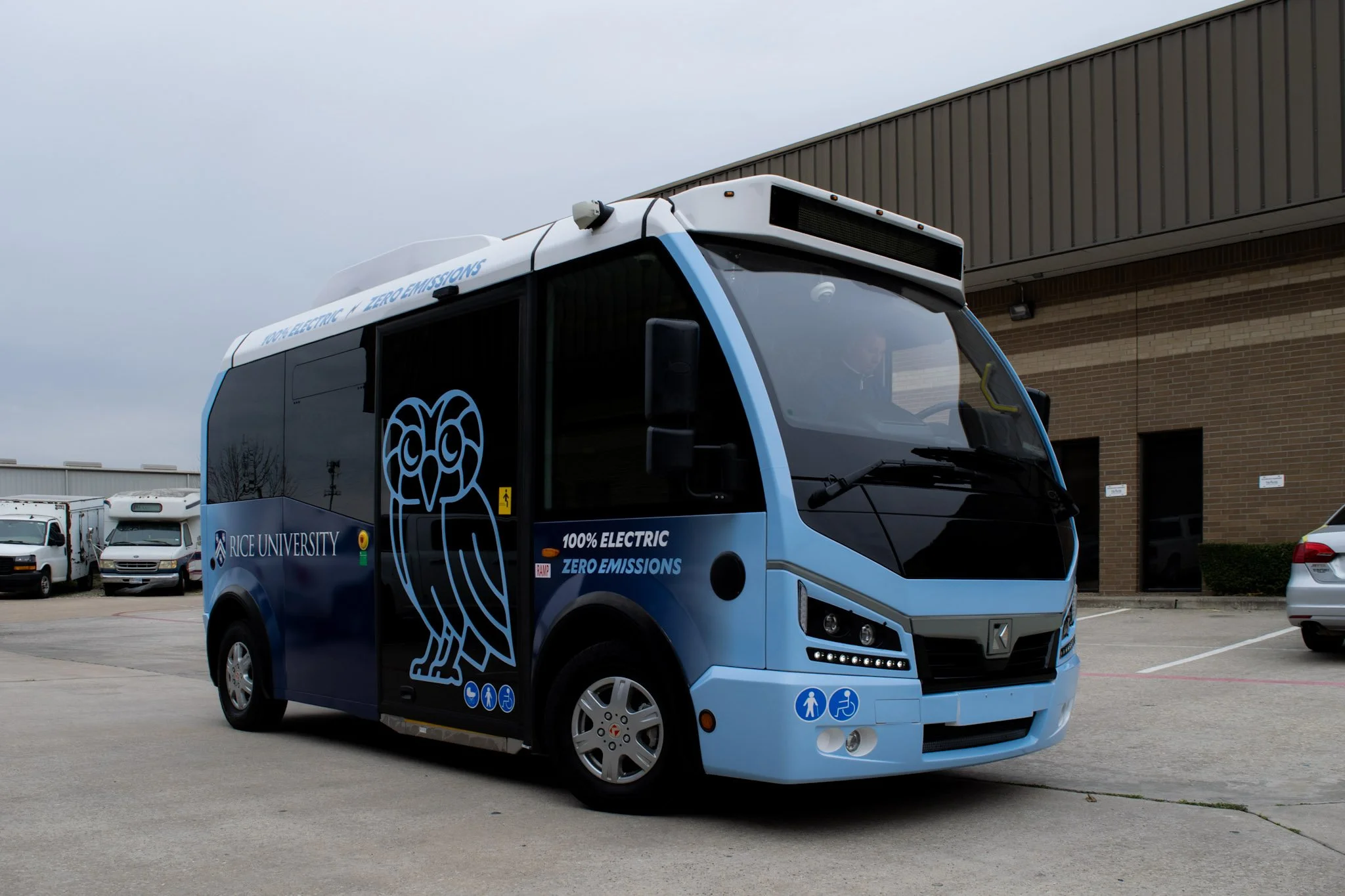 Electric Rice University shuttle bus with a custom vehicle wrap parked outside a commercial building in Houston, Texas.