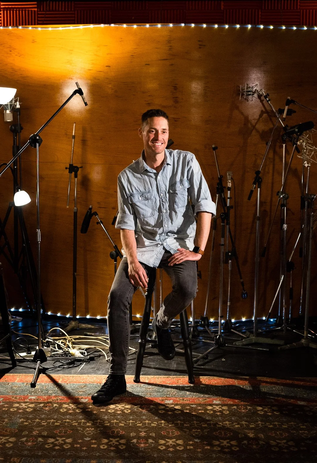 Man sitting on a stool in recording studio surrounded by microphones and audio equipment, smiling at camera.