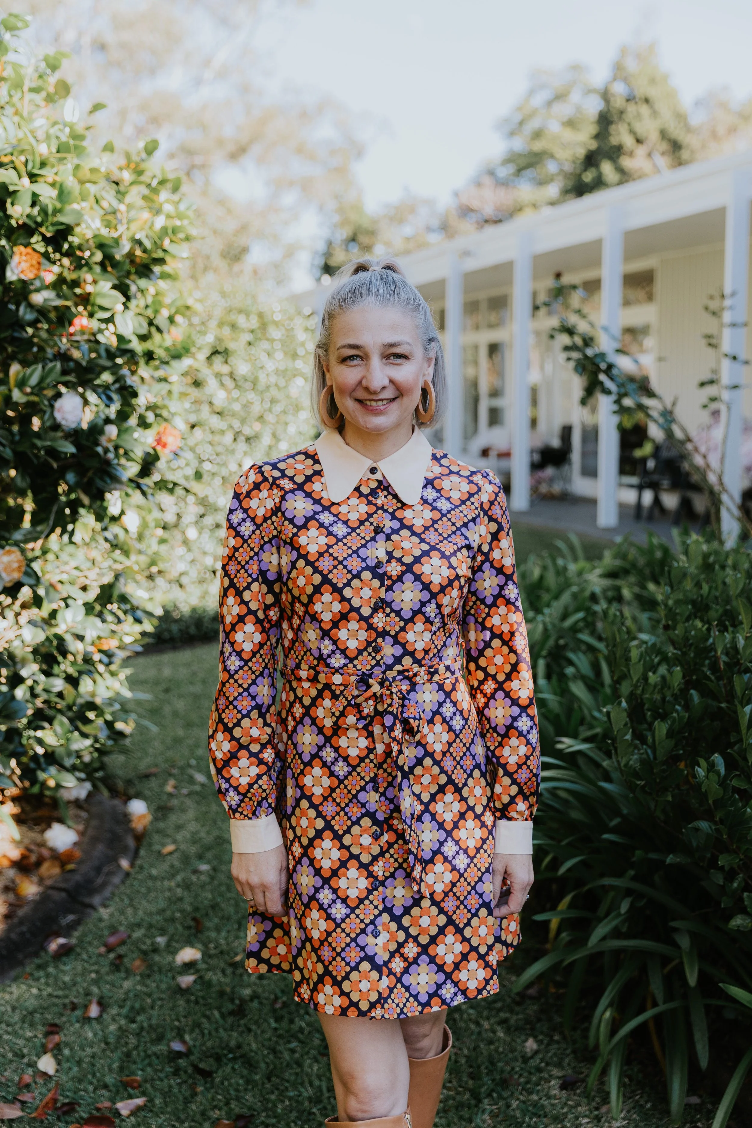 A woman standing outdoors, smiling, wearing a colorful, patterned dress with a white collar and cuffs, and tan boots, in a garden with green bushes and trees, and a white house in the background.
