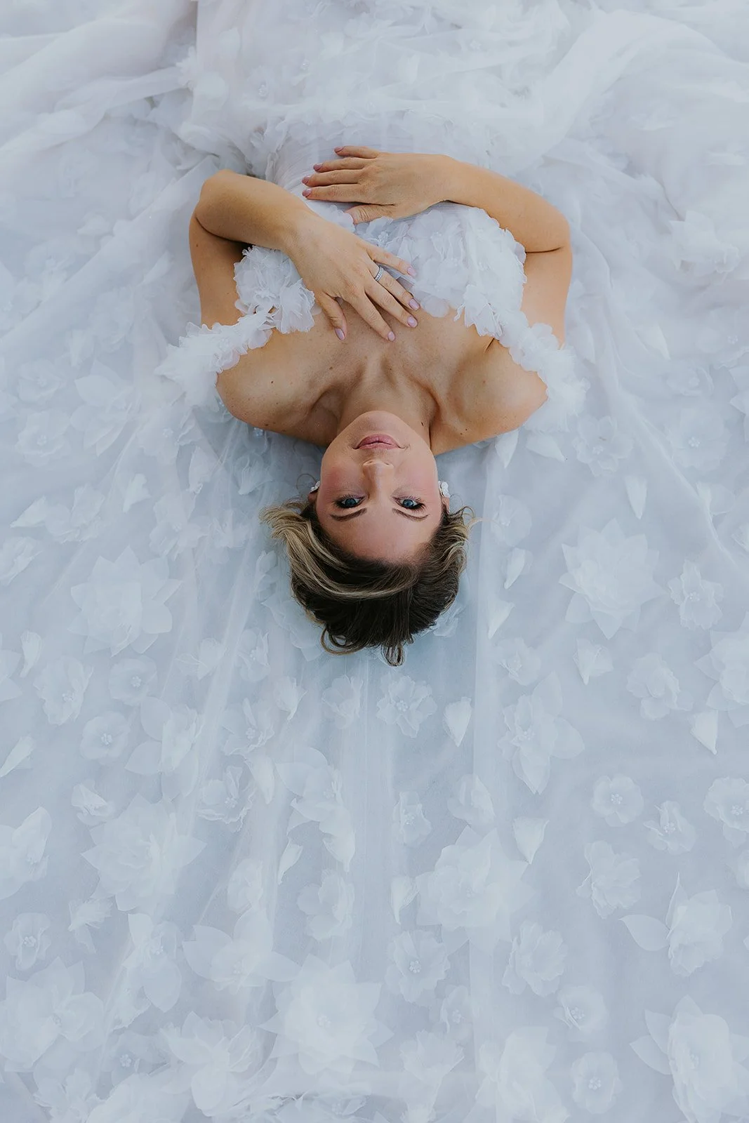A woman in a white wedding gown lying on her back on a fabric with floral details, looking up at the camera.