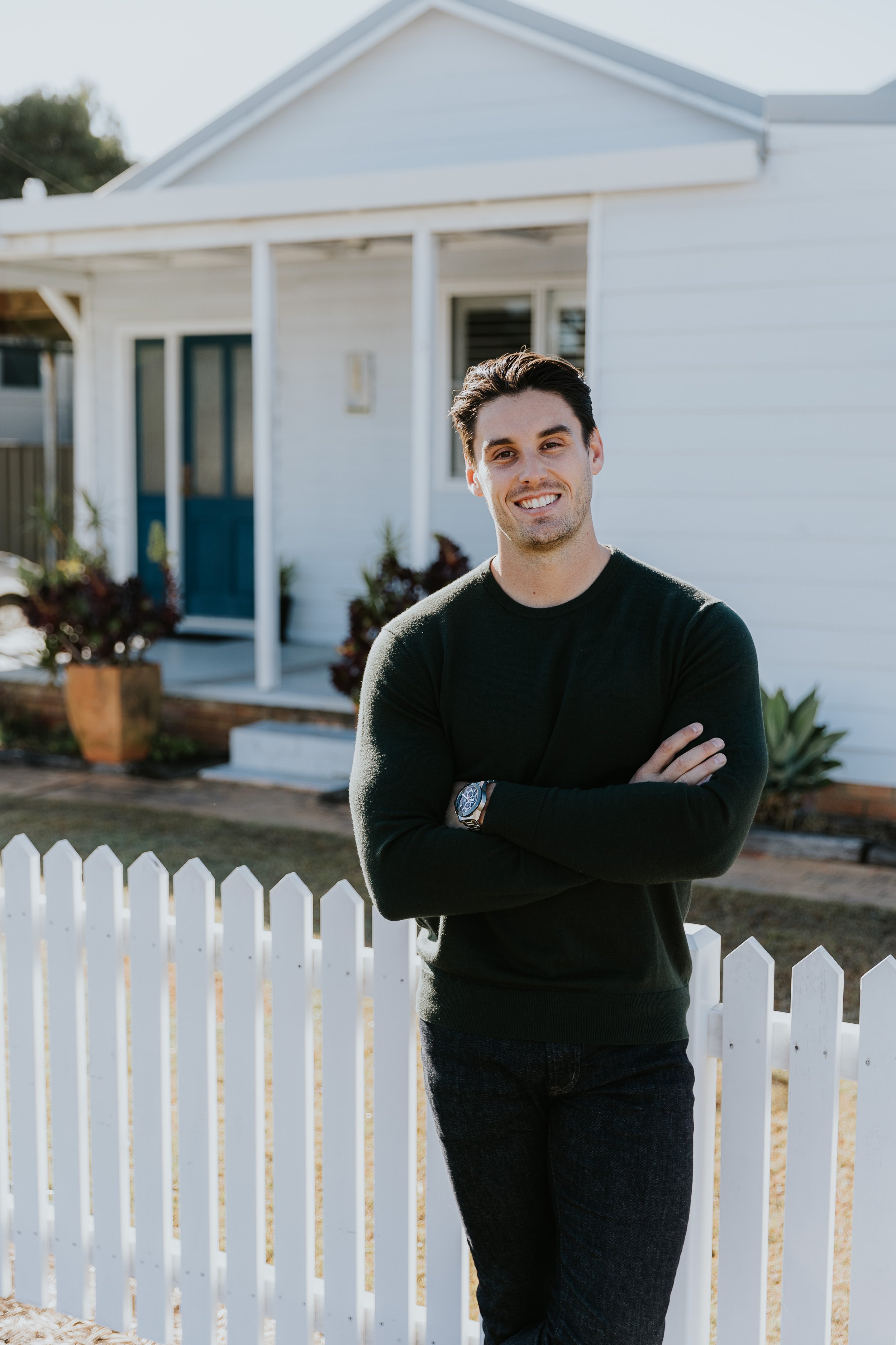 A young man with dark hair, wearing a black sweater and jeans, stands with arms crossed in front of a white house with a picket fence, smiling at the camera.