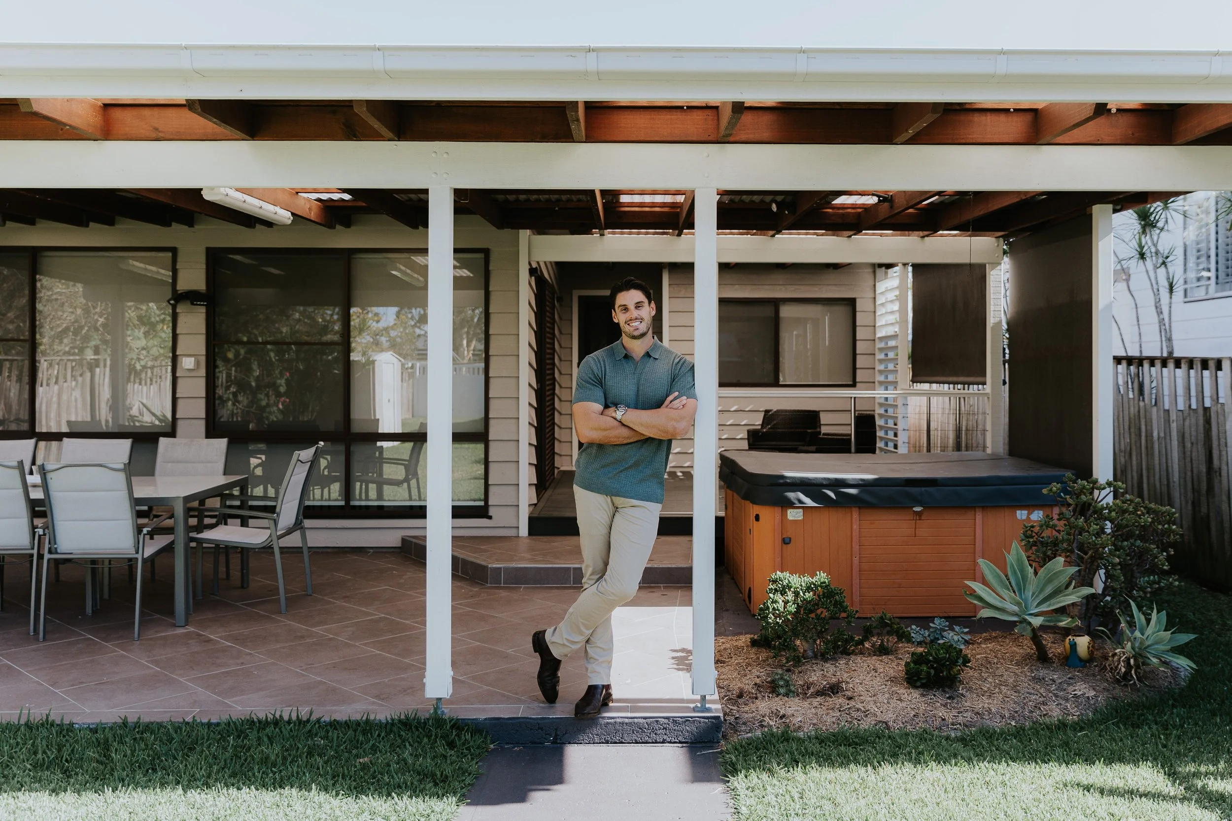 Young man standing outside on porch of house, smiling with arms crossed, next to hot tub, patio table and chairs, plants, and backyard fencing.