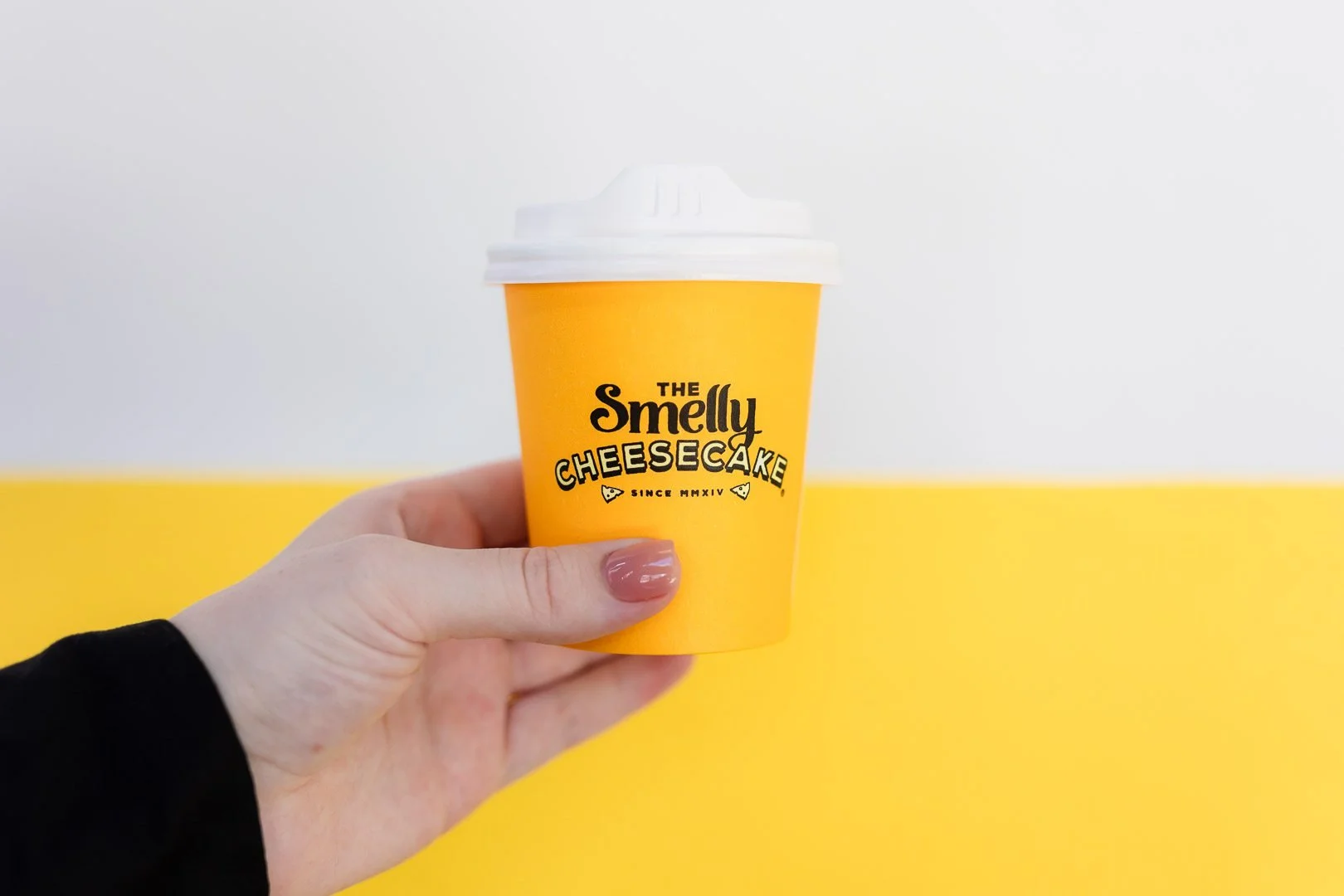 A person holding a yellow to-go coffee cup with a white lid, logo reading 'The Smelly Cheesecake' on a yellow and white background.