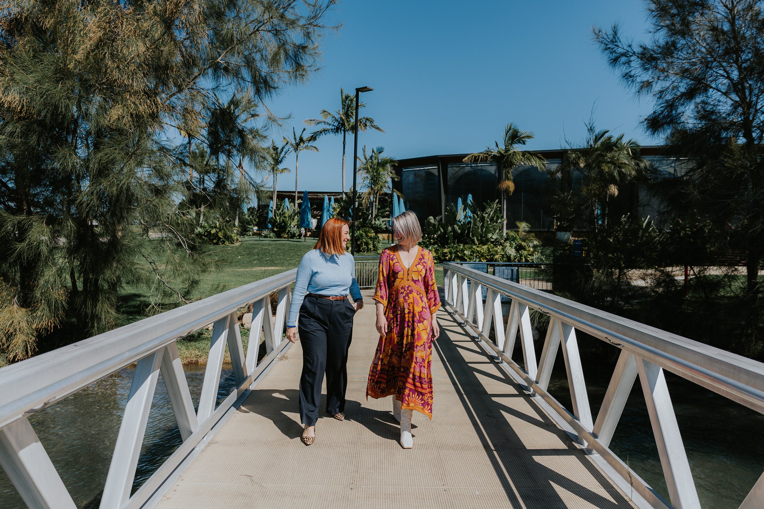 Two women walking and talking on a bridge in a lush outdoor park area with trees, grass, and blue umbrellas in the background.