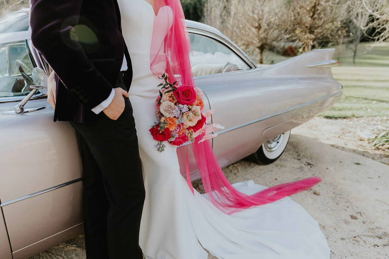 A bride and groom in wedding attire standing next to a vintage car decorated with pink and peach flowers and pink tulle ribbons.