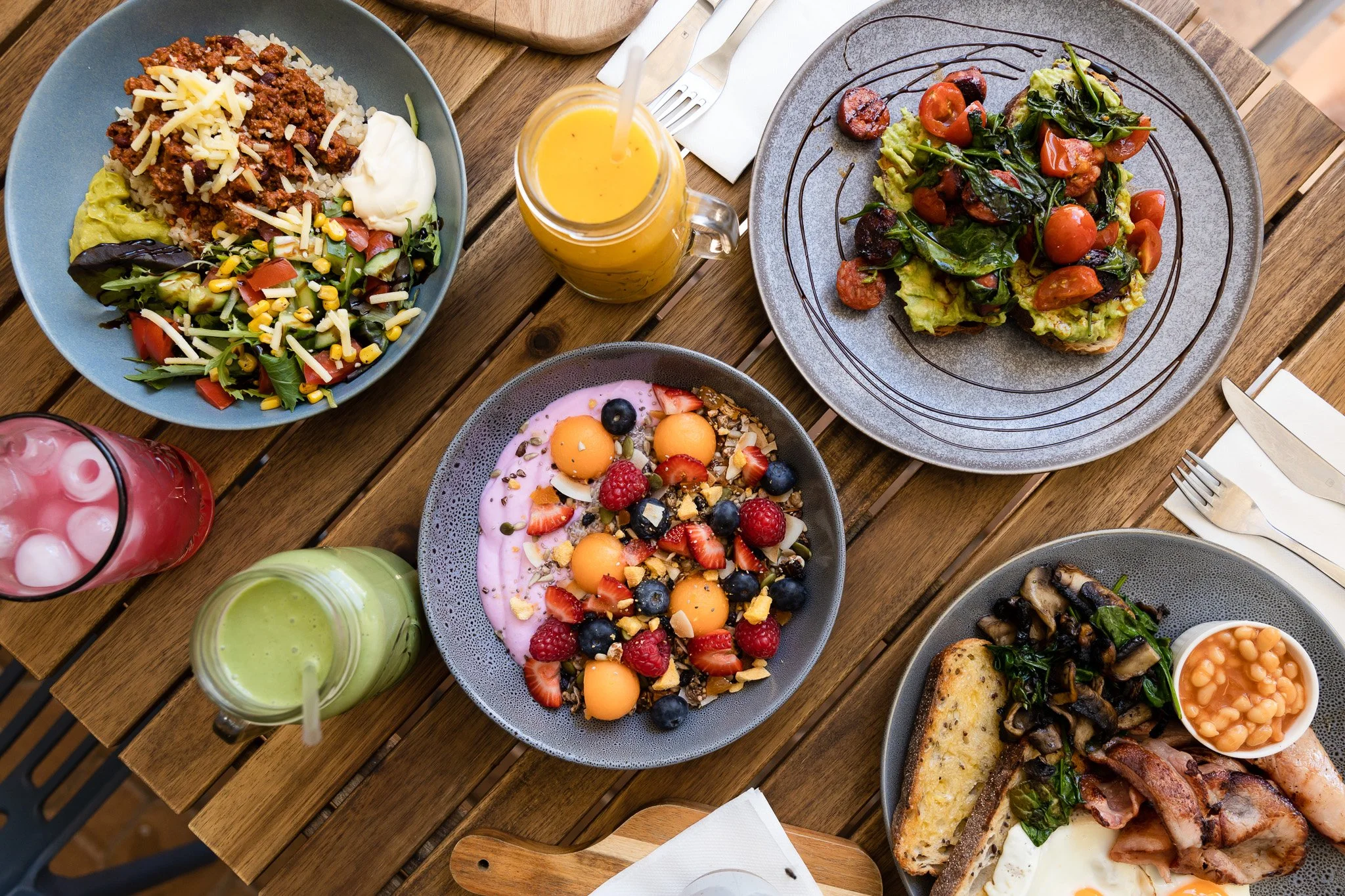 A variety of breakfast dishes on a wooden table including a bowl of rice, ground beef, and vegetables, a plate of toast with tomatoes and greens, a bowl of mixed berries and fruit, another drink with strawberries, and a plate with baked eggs, bacon, 