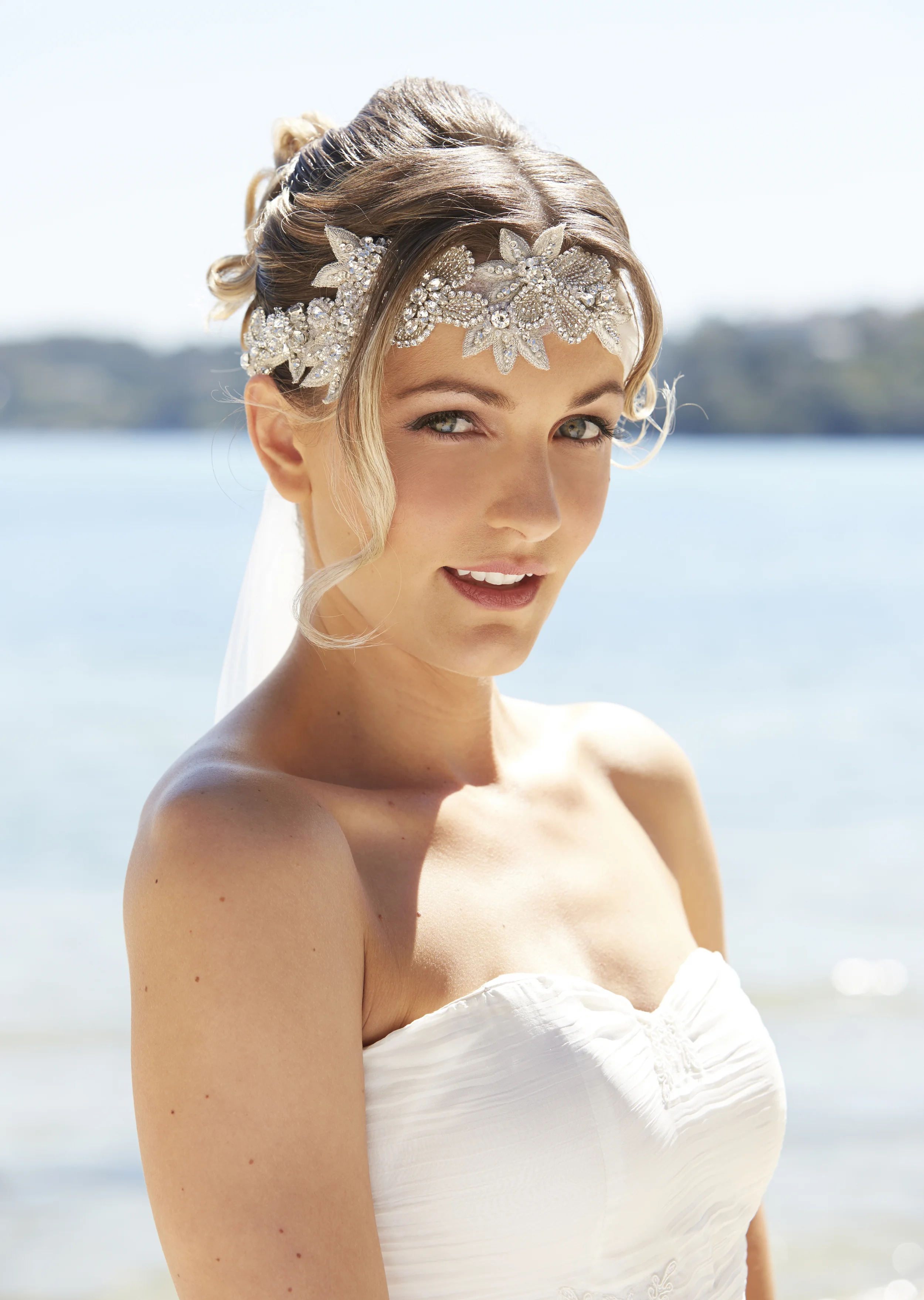 A woman wearing a strapless white dress and a jeweled floral headpiece, standing outdoors near water with a blurred shoreline in the background.