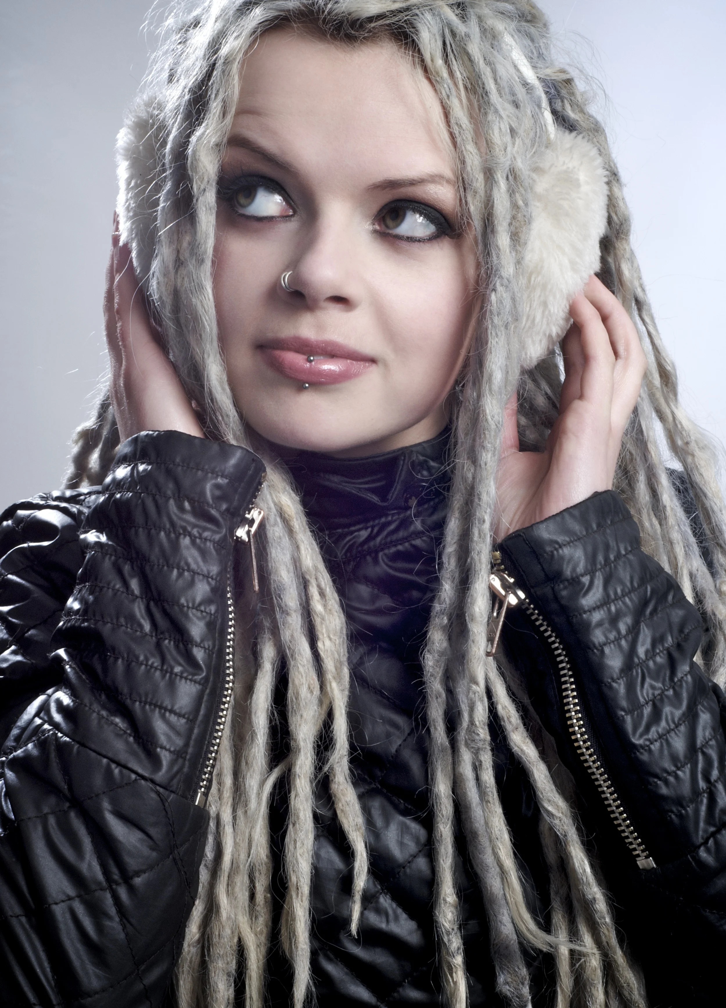 A young woman with blonde dreadlocks, wearing a black leather jacket and fluffy earmuffs, listening to music with her eyes looking upwards.
