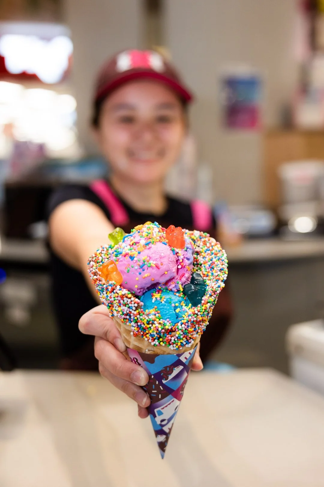 Person holding a colorful ice cream cone with sprinkles and gummy candies, with a blurred smiling person in the background.