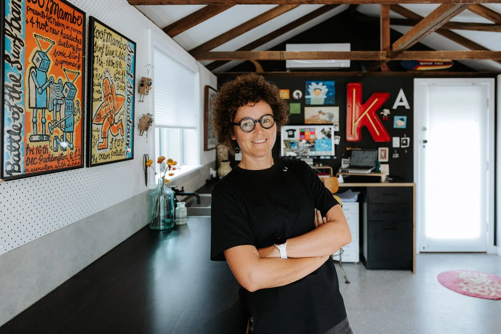 Sydney Portrait Photography for Graphic Design small business. A smiling woman with curly hair and glasses, standing with arms crossed inside a creative workspace with colorful artwork and a large red letter K on the black wall.