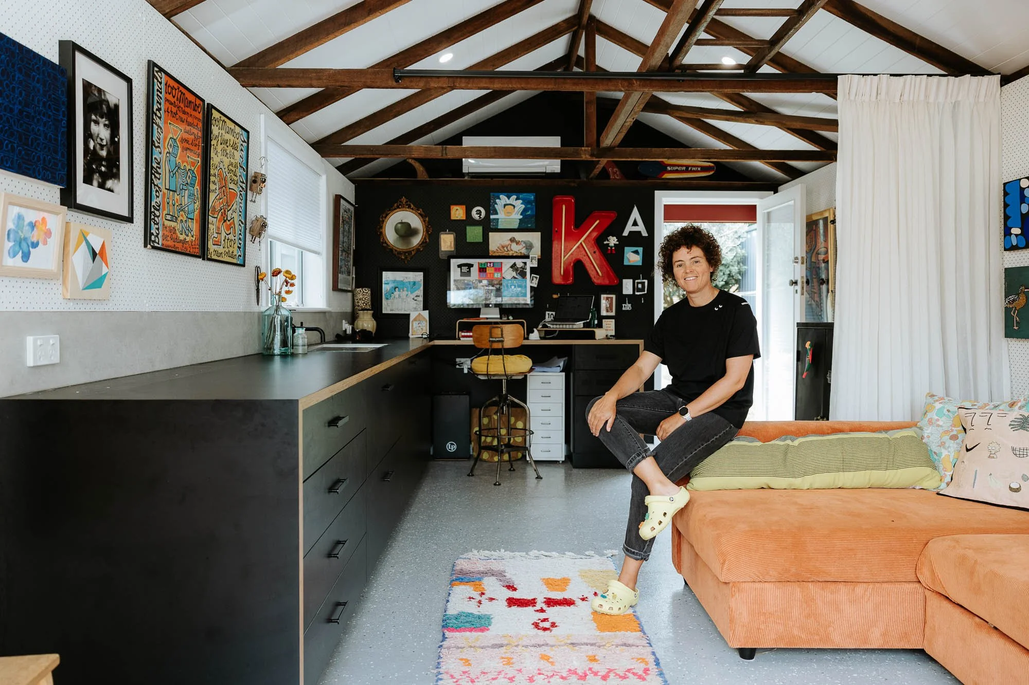 Woman sitting on an orange sectional sofa in a colorful, art-filled living room with a kitchen area in the background. She wears a black t-shirt, gray jeans, and yellow Crocs, smiling at the camera.