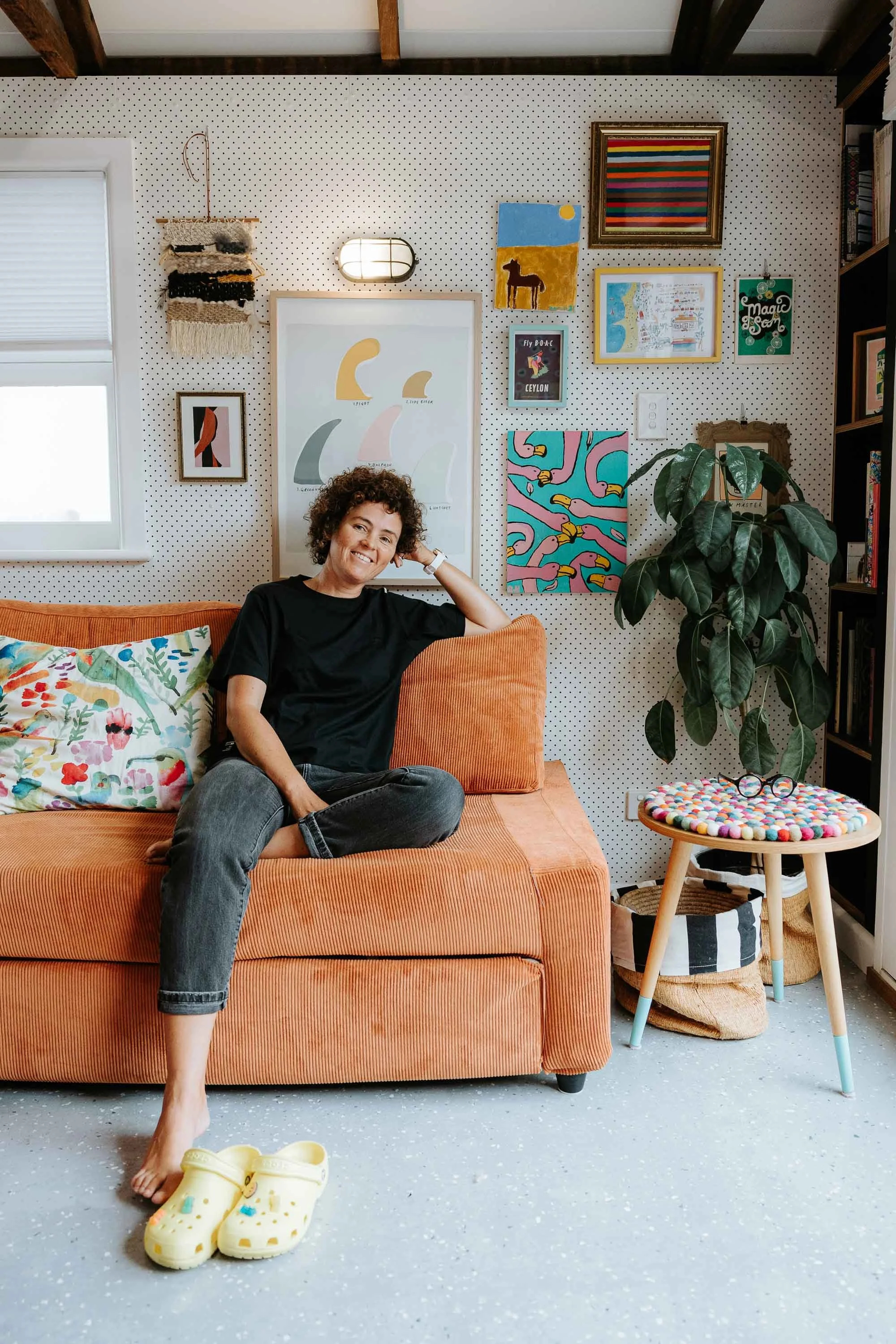 A woman sitting on an orange couch in a colorful, creatively decorated room with art on the walls, a large green plant, a small round table with a multicolored pom-pom cover, and a pair of yellow Crocs slippers on the floor.