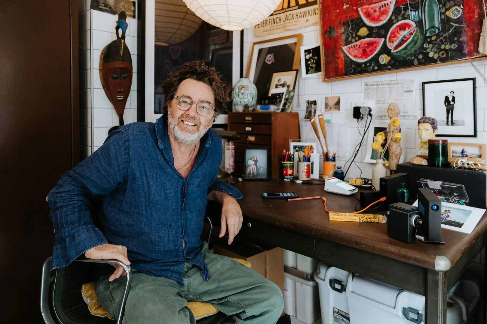 A man with glasses, curly hair, and a beard smiling and sitting at a cluttered art studio desk with sculptures, framed photos, and art supplies surrounded by colorful artwork and decorations.