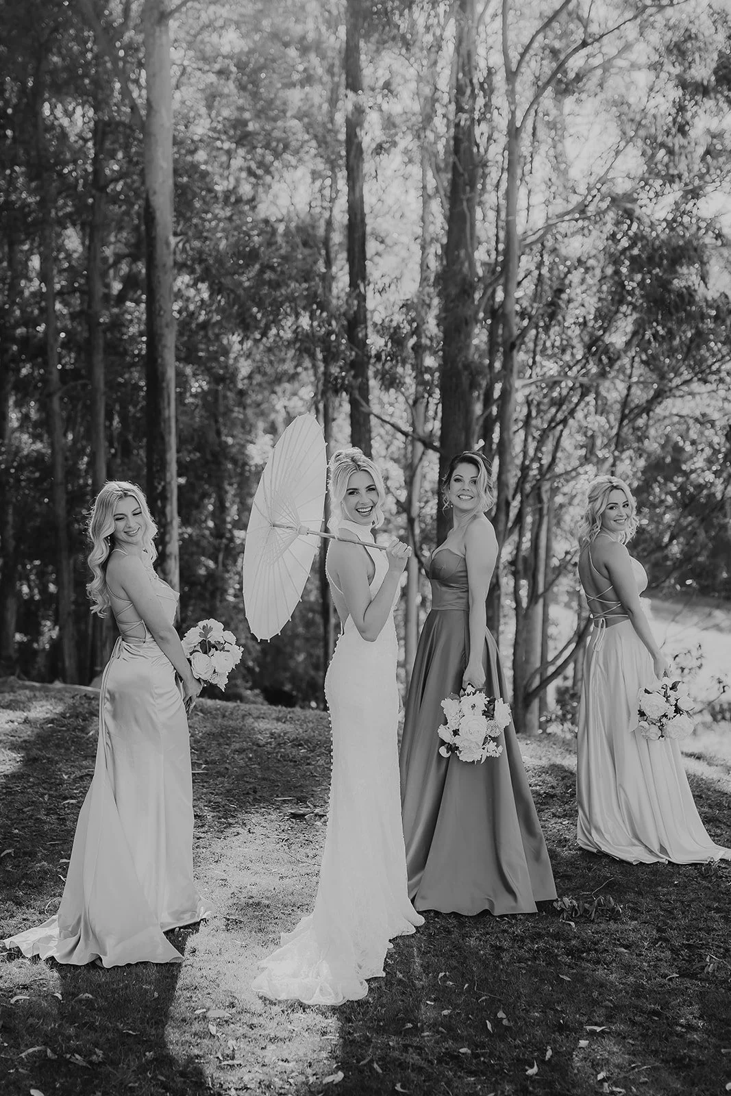 Four women in wedding dresses and bridesmaid dresses outdoors in a forested area, holding bouquets, with one woman holding a parasol, all smiling.