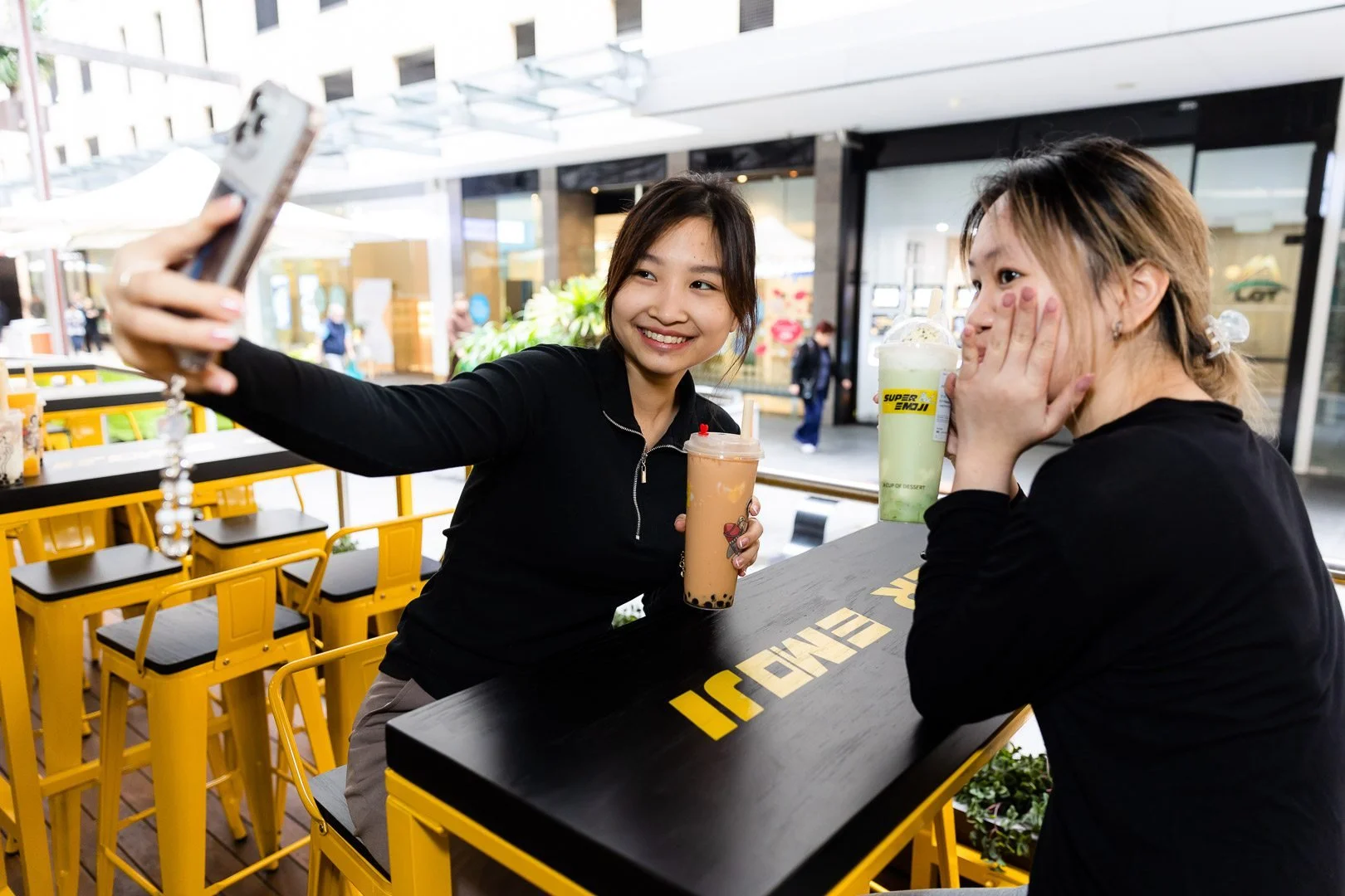 Two young women sitting at an outdoor cafe table with bright yellow chairs, taking a selfie. One is smiling and holding a bubble tea, the other is covering her face with her hands, both casually dressed.