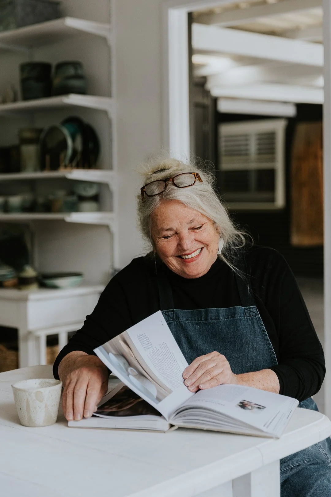 An elderly woman with white hair and glasses on her head, smiling while looking at a cookbook in a kitchen.