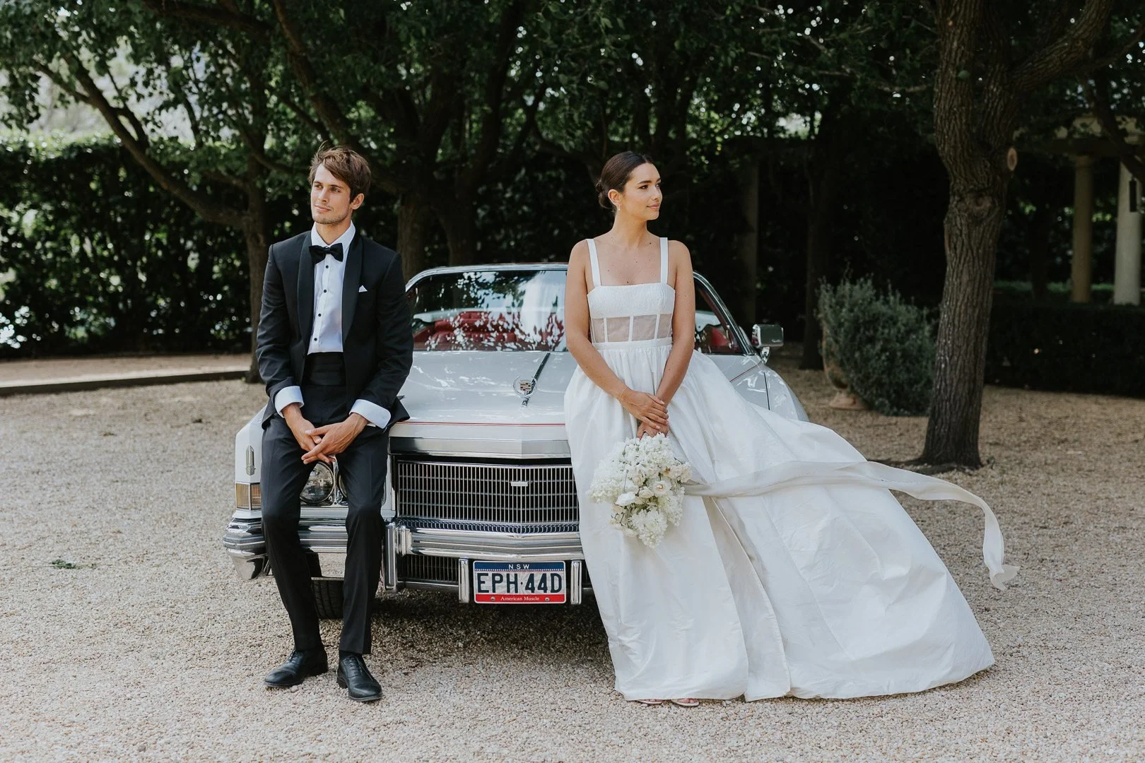 A bride and groom in wedding attire outdoors near a vintage car, with trees and greenery in the background. The groom is sitting on the front of the car, and the bride is standing beside it, holding a bouquet of white flowers.