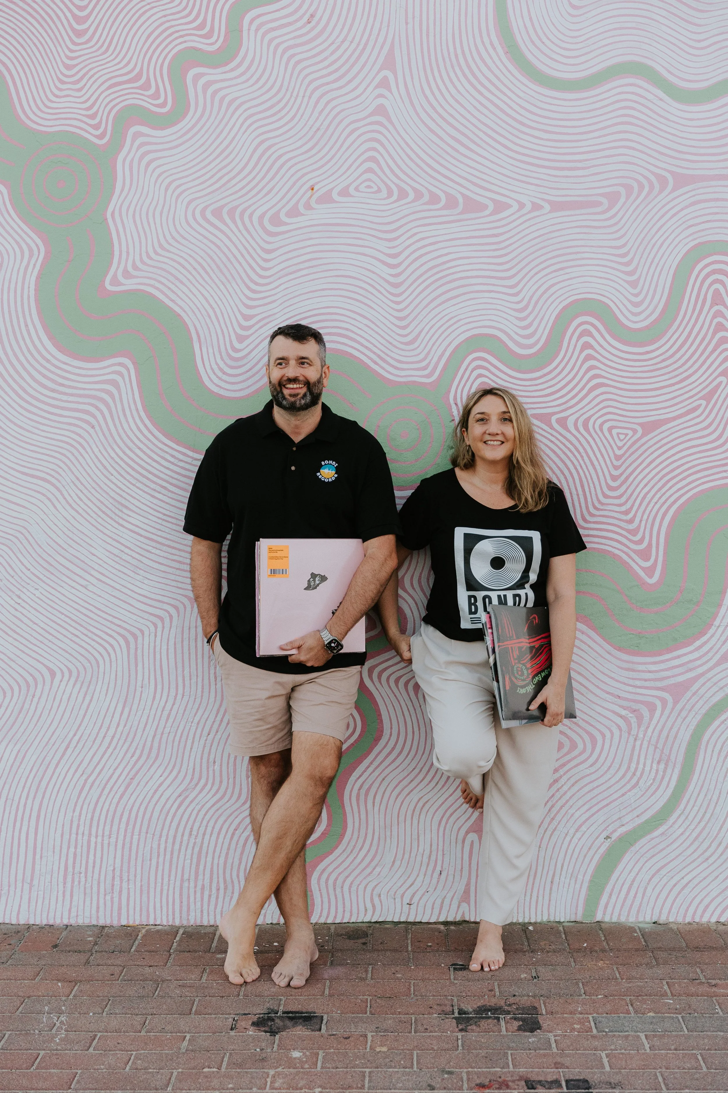 A smiling man and woman standing barefoot against a colorful, abstract mural, holding vinyl records.