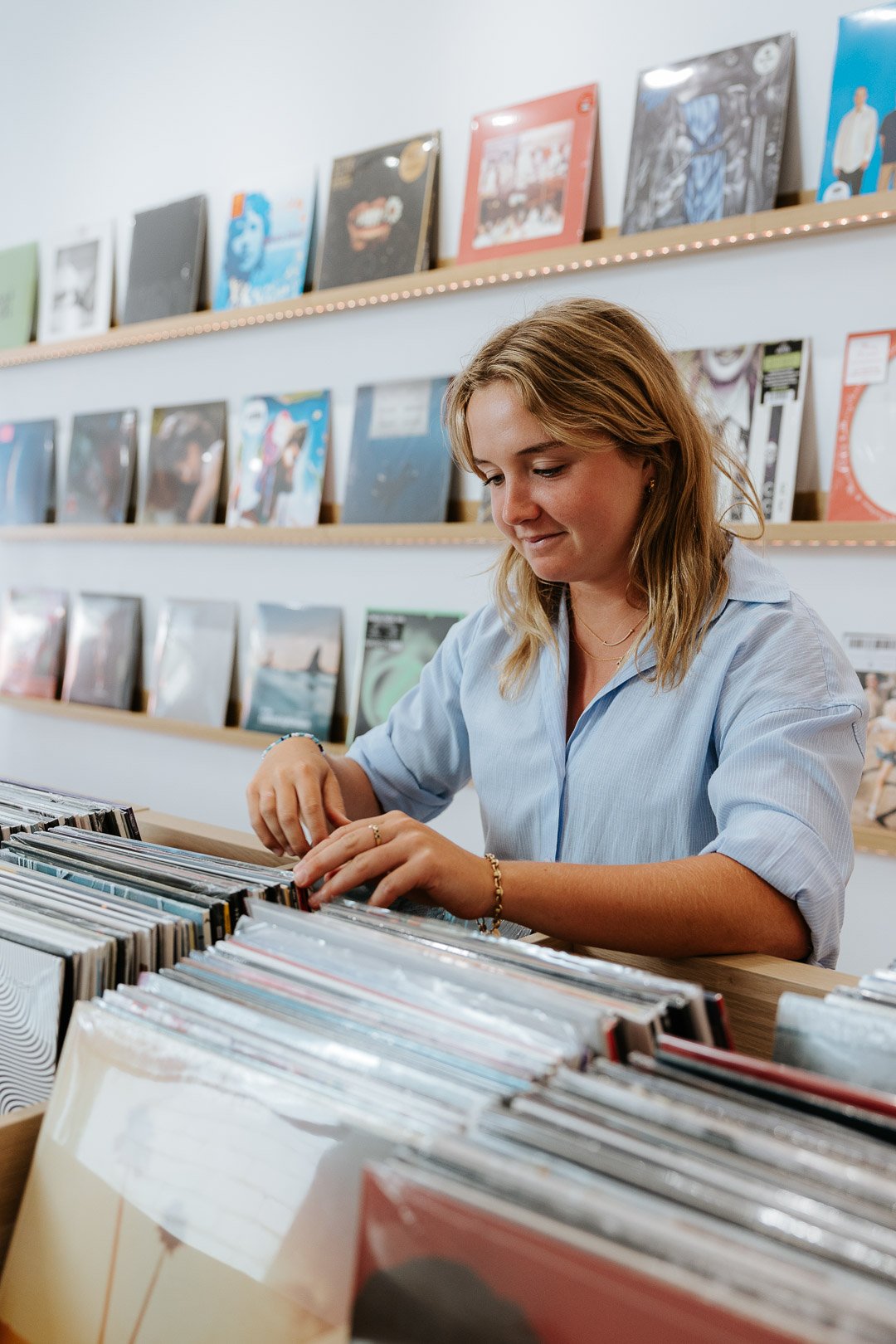 A young woman browsing through records or vinyl albums at a record store, with shelves of colorful album covers in the background.