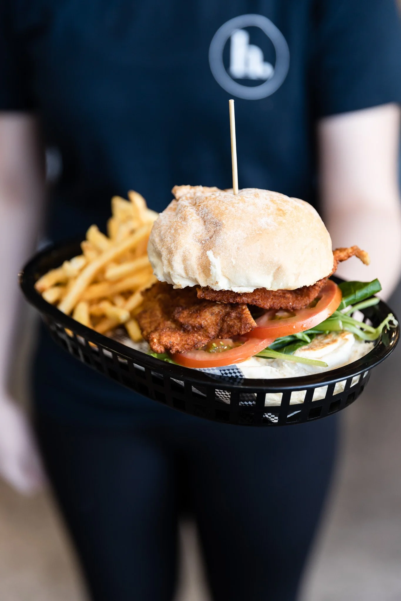 Close-up of a serving basket with a fried chicken sandwich, French fries, and pickles, held by a server wearing a dark uniform.