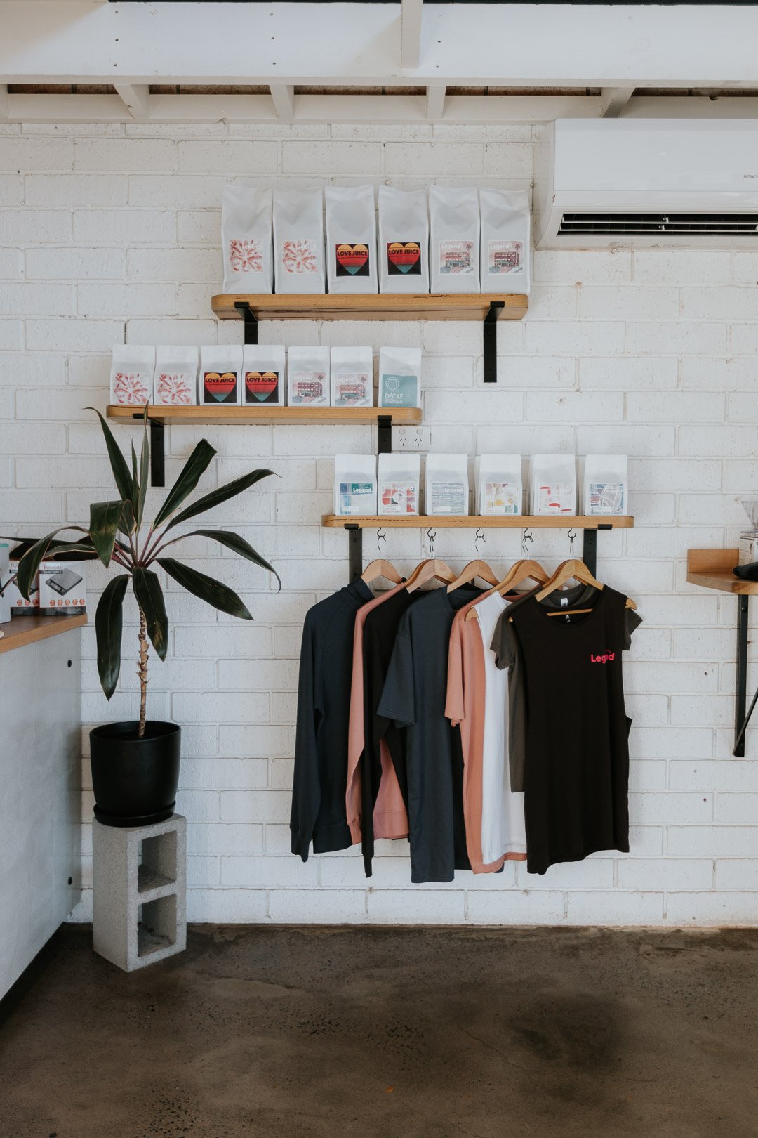 Shop interior with shelves holding coffee bags, a potted plant on cinder block, and hanging clothes on a wooden and metal rack.