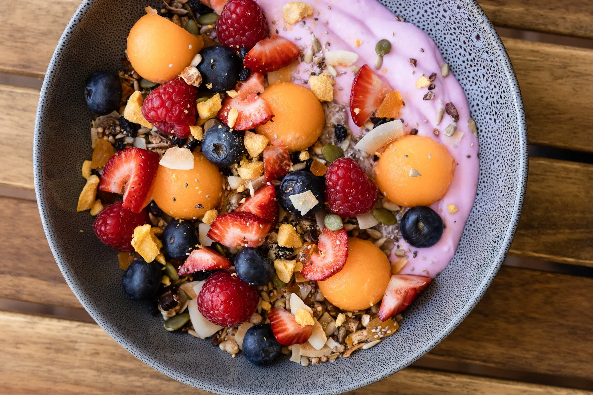 A bowl of yogurt topped with fresh strawberries, raspberries, blueberries, sliced melons, granola, seeds, and crumbled cookies on a wooden table.