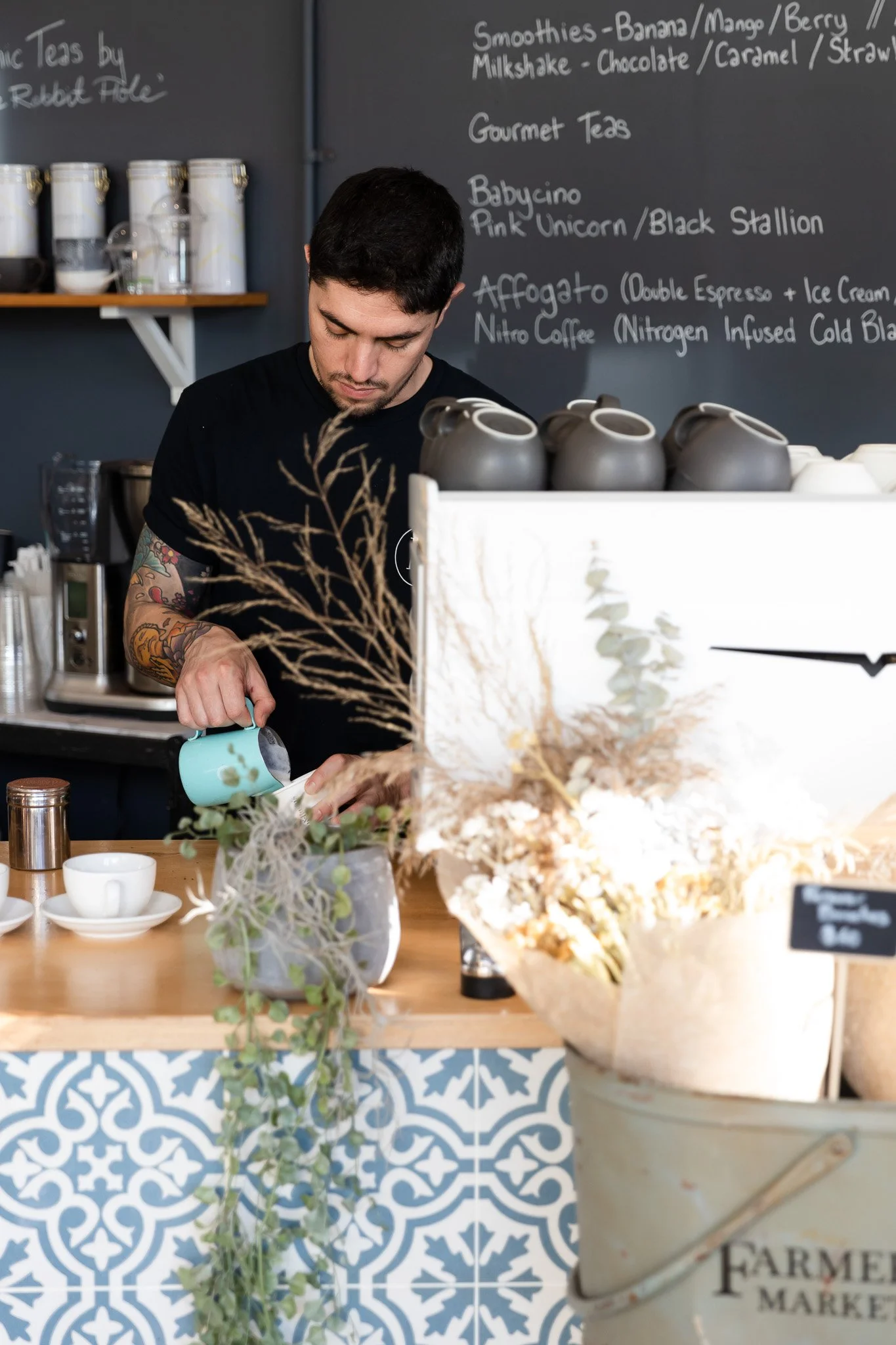 Barista pouring milk or cream with a pitcher at a coffee shop counter, with a menu board in the background listing smoothies, teas, and specialty coffee drinks.