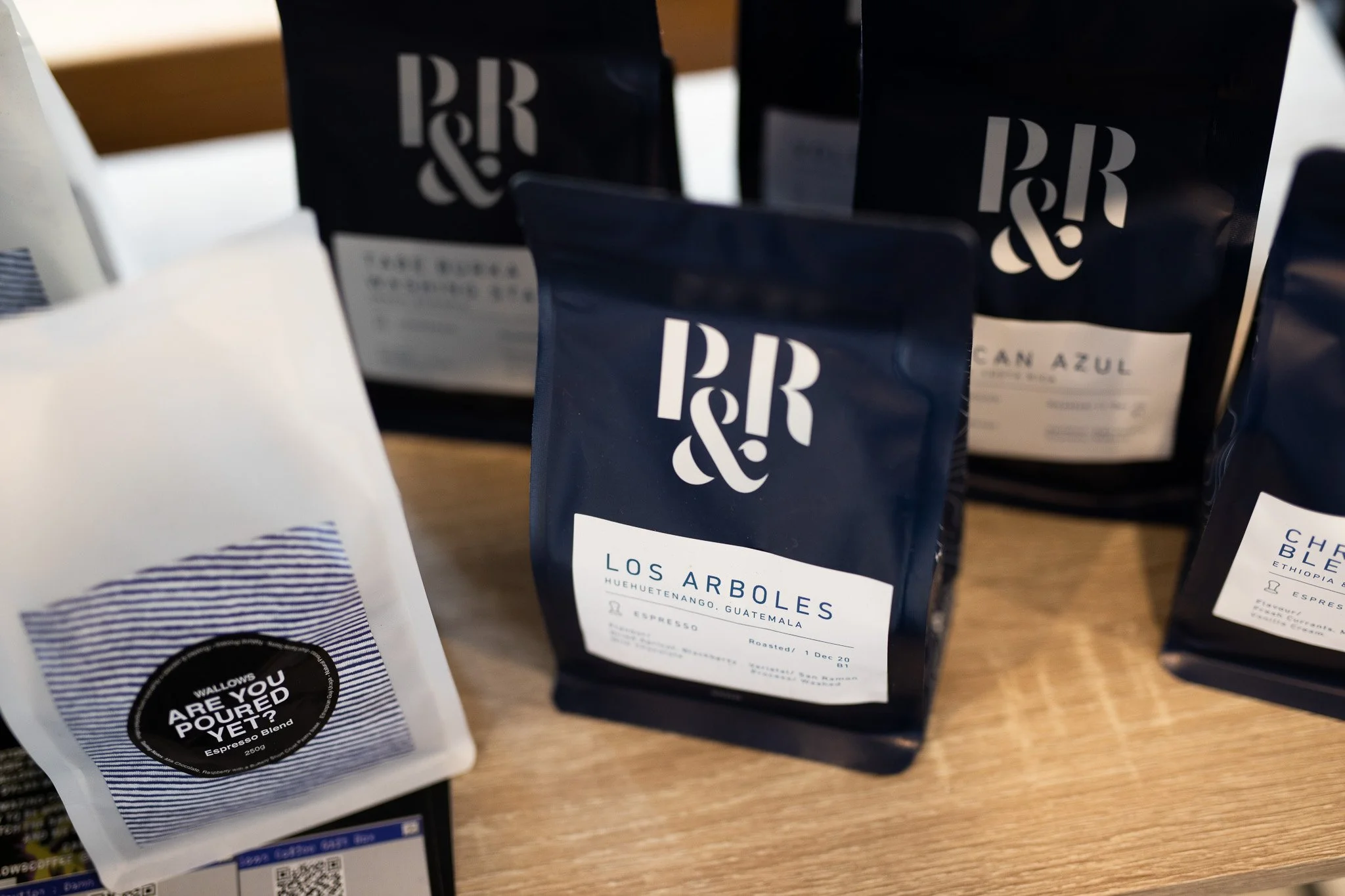 Several bags of coffee beans on a wooden surface, with labels displaying different coffee varieties and roaster details.