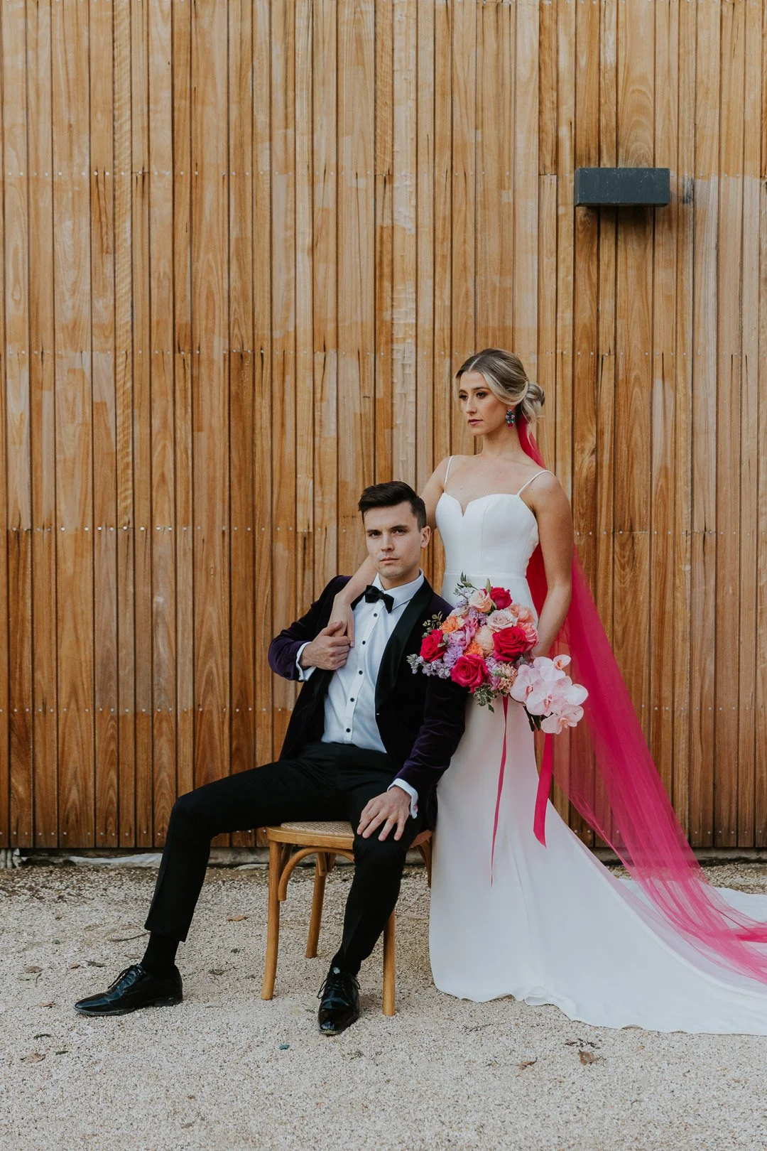 A bride and groom in wedding attire posing outdoors in front of a wooden wall. The bride wears a white dress with a long pink veil and holds a bouquet of red, pink, and white flowers. The groom sits on a chair in a tuxedo, with one hand on his lap an