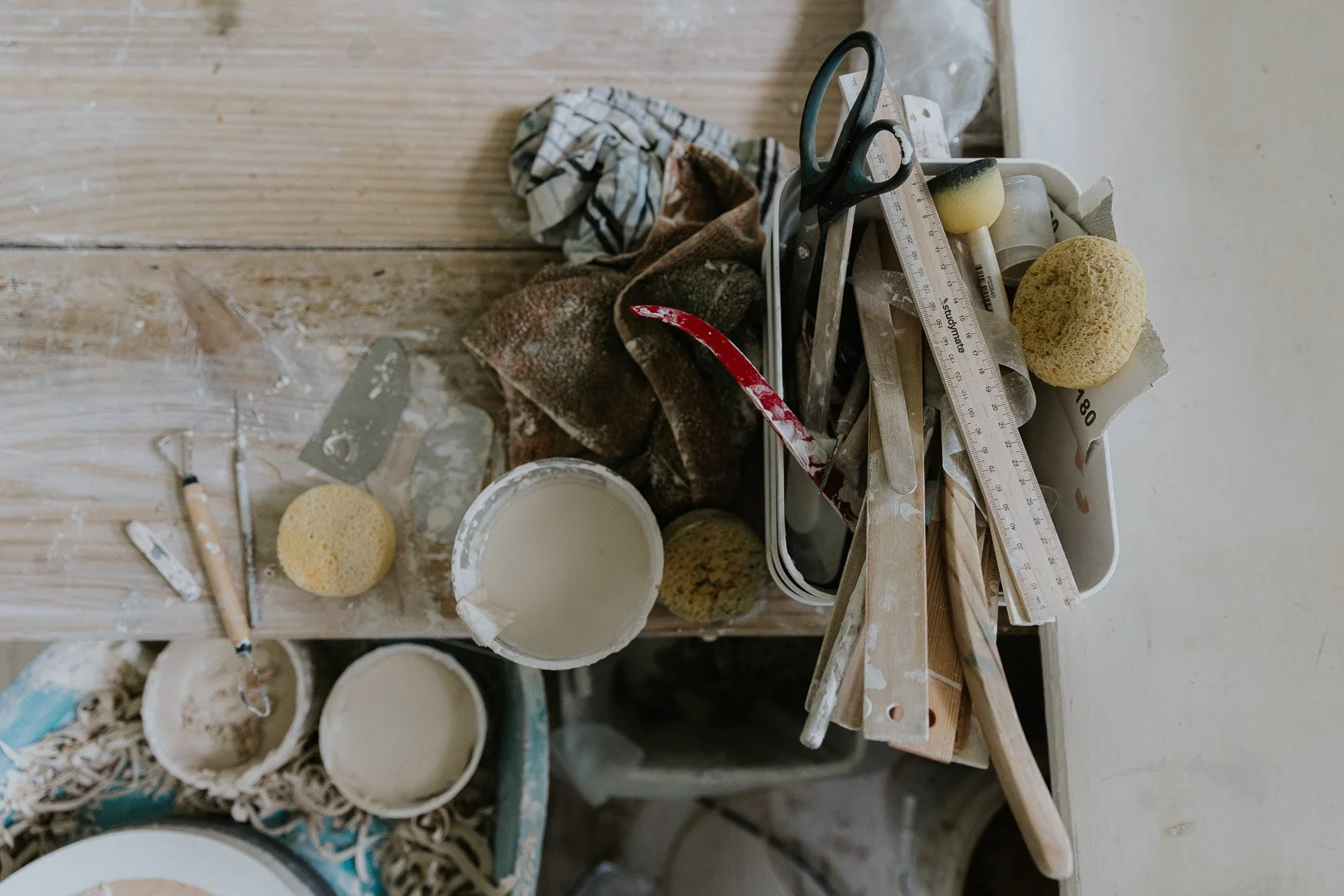 A cluttered workspace with drywall tools, sponges, buckets of white paint, a measuring ruler, scissors, and paint supplies on a table.