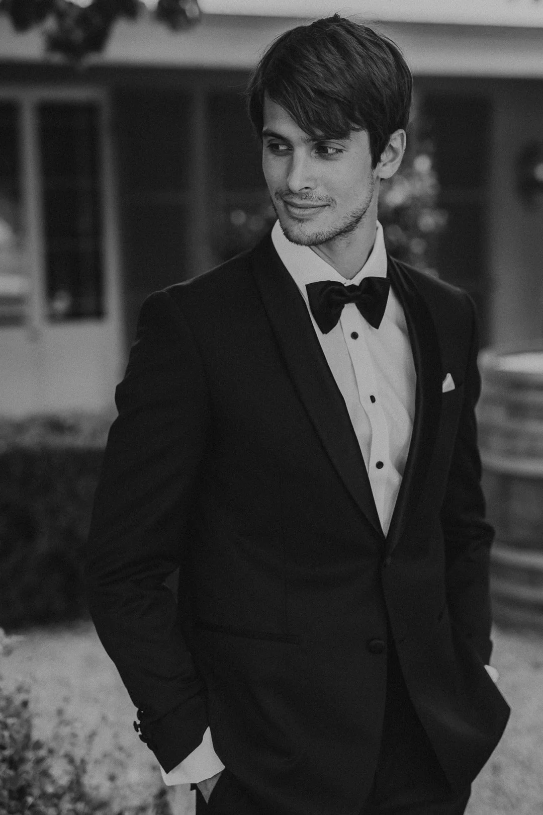 A young man in a tuxedo with a bow tie, standing outdoors at night with hands in pockets.