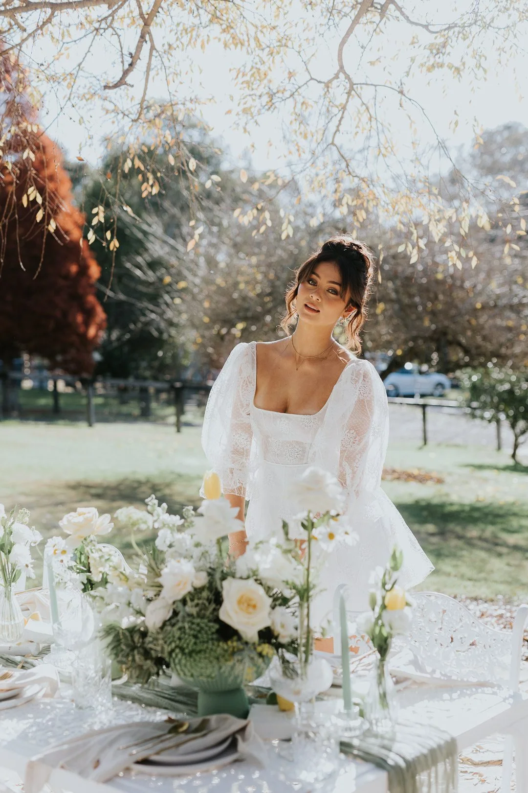 A young woman in a white lace dress standing behind a table decorated with white flowers outdoors on a sunny day.