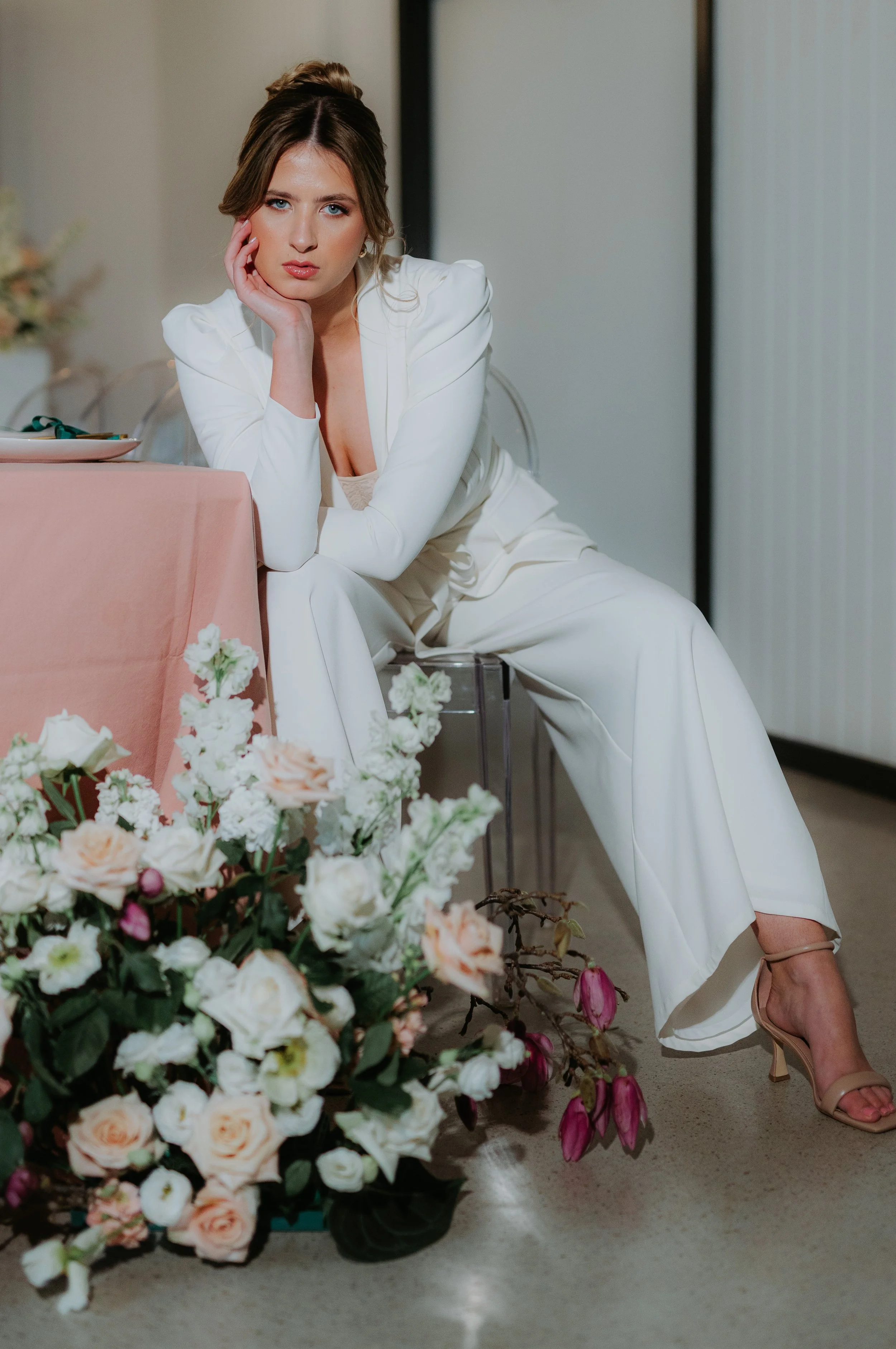 A woman in a white pantsuit sitting at a table with a floral arrangement in front of her, looking at the camera with her hand resting on her face.