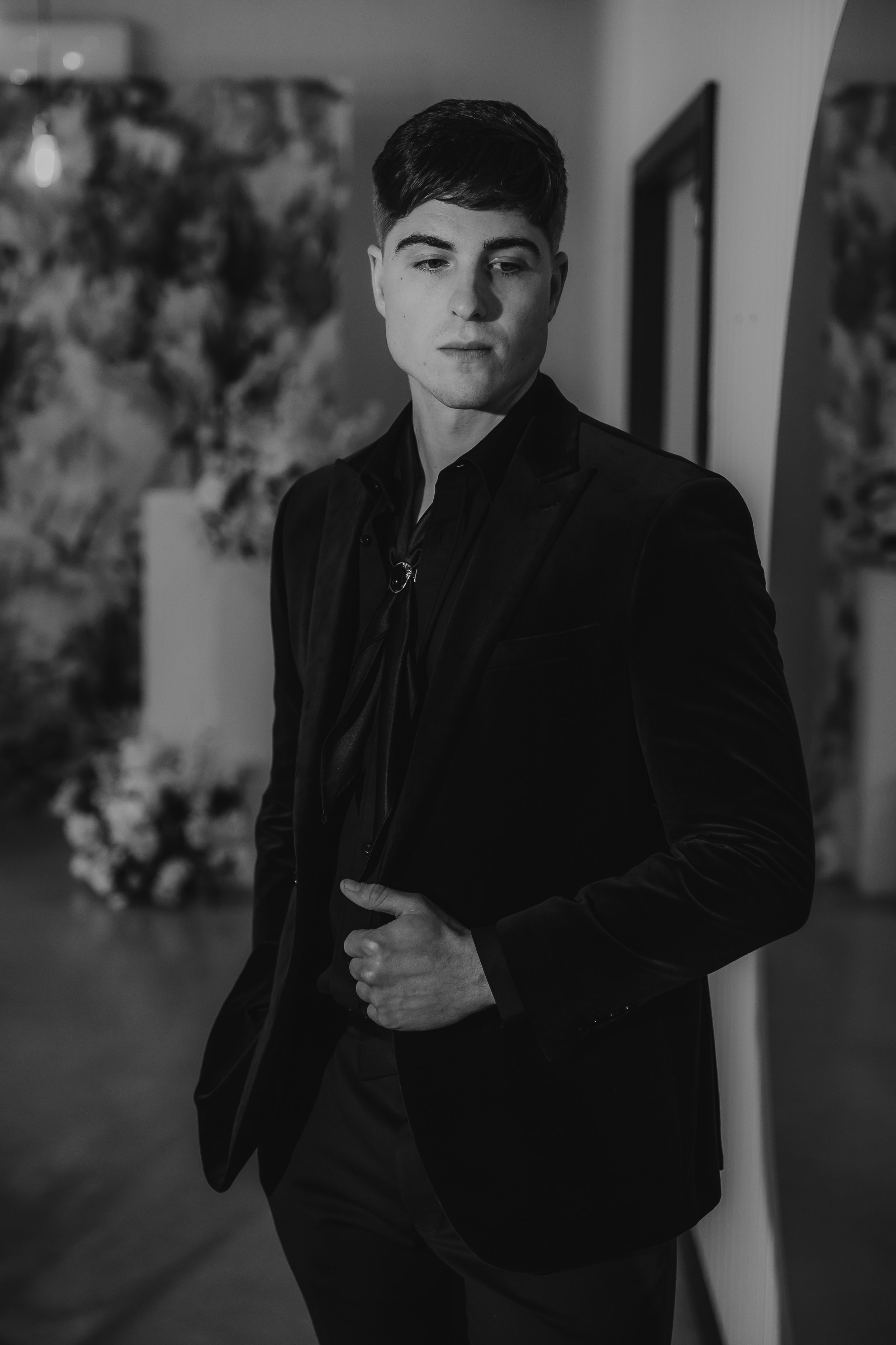 A young man in a black tuxedo standing indoors, looking off to the side with his hand on his stomach, with blurred artwork or decorations in the background.